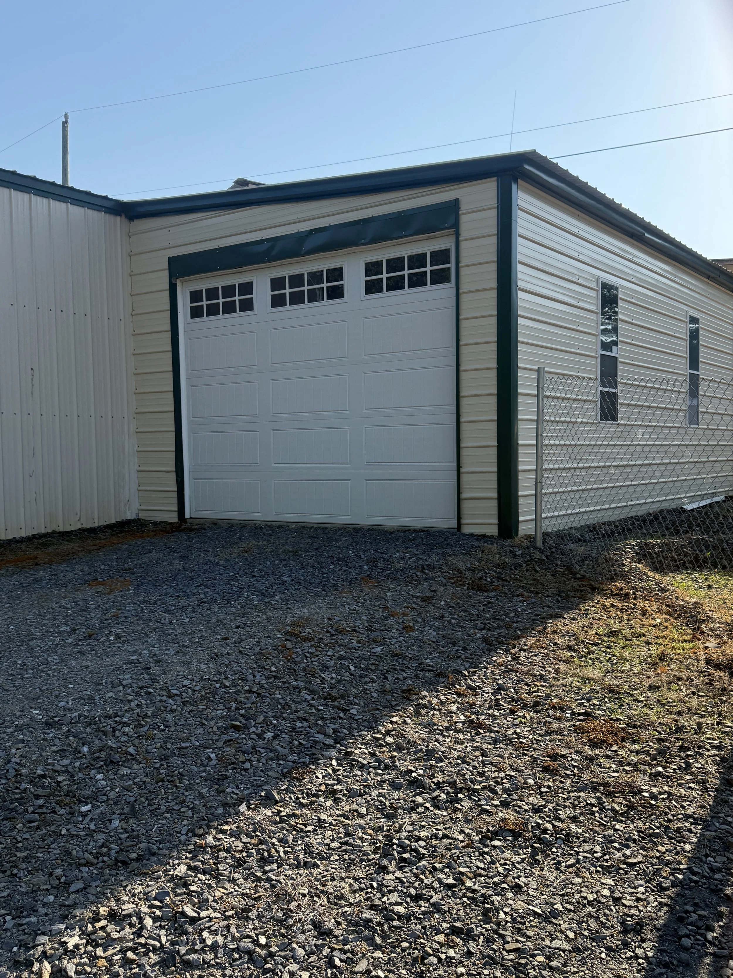A large white garage door on a beige metal building next to a chain-link fence, with two narrow vertical windows and a gravel ground, under a clear blue sky.