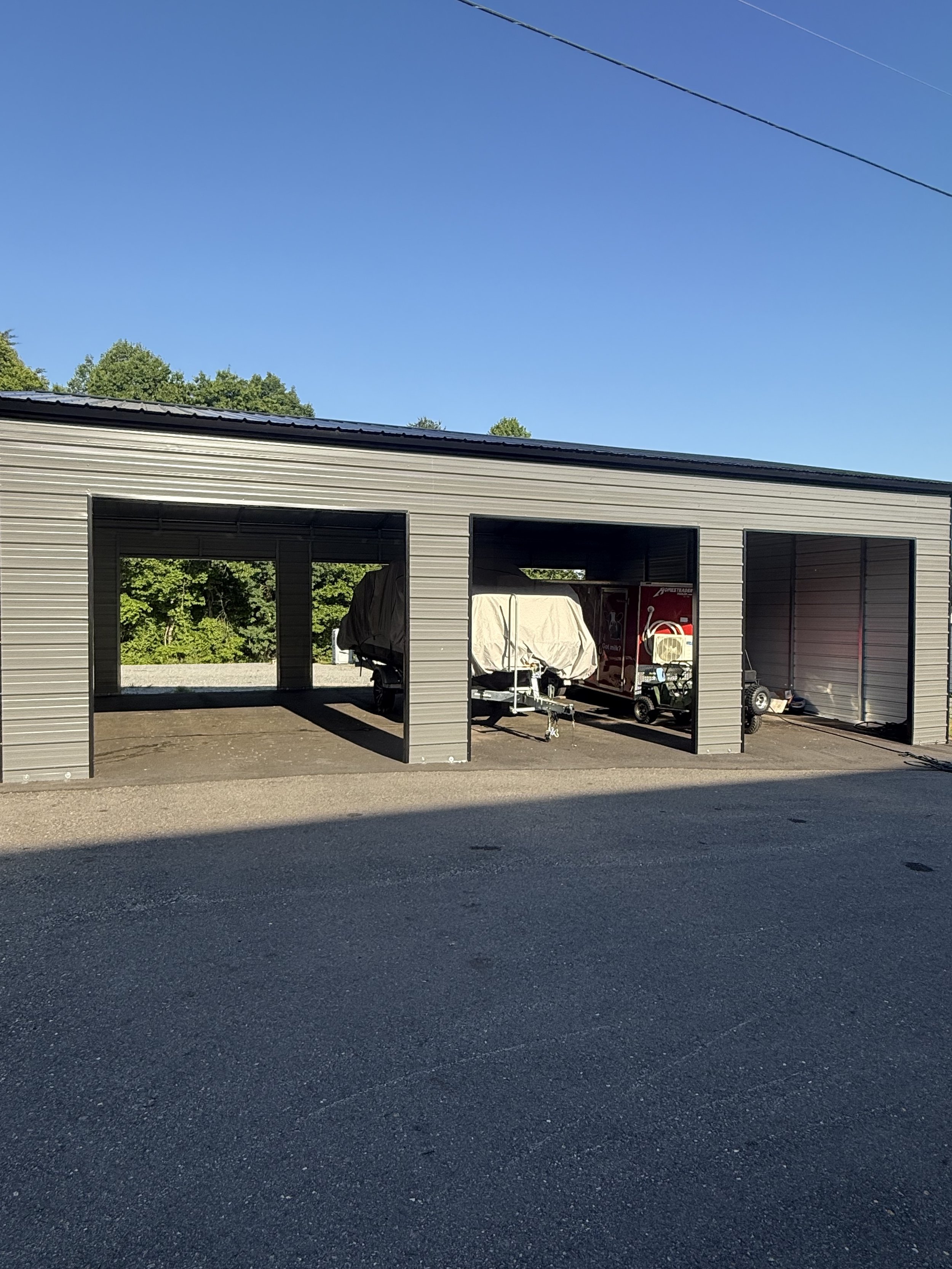 A metal storage building with open bays, a covered boat, a red wagon, and a golf cart inside, on a paved surface with trees and a clear blue sky in the background.