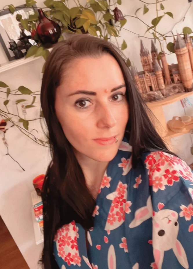 Eine junge Frau mit dunklem, langem Haar trägt einen Kimono mit Hasen- und Kirschblütenmuster. Im Hintergrund sind Pflanzen und Dekorationsgegenstände zu sehen.