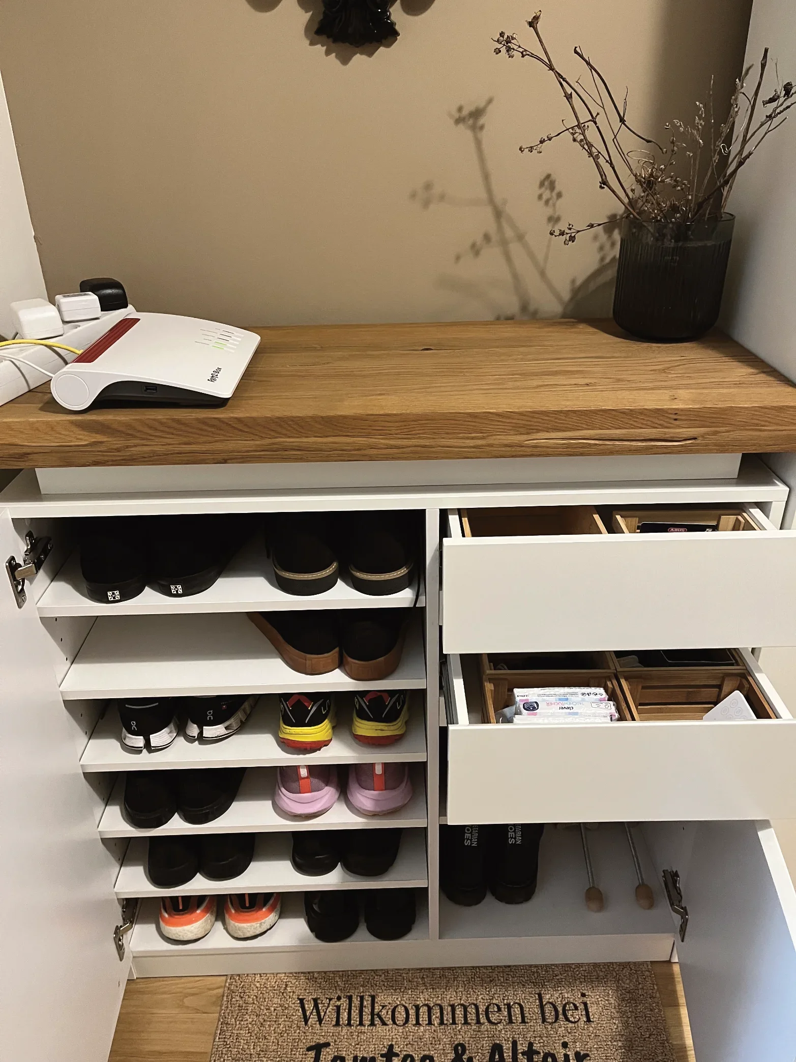 Ein Schuhschrank mit mehreren Regalen voll mit verschiedenen Schuhen, darunter Sneakers, Sportschuhe und Hausschuhe, auf einem Holzboden. Im Vordergrund liegt ein Begrüßungsmatten mit dem Text 'Willkommen bei Tamtam & Alina'.