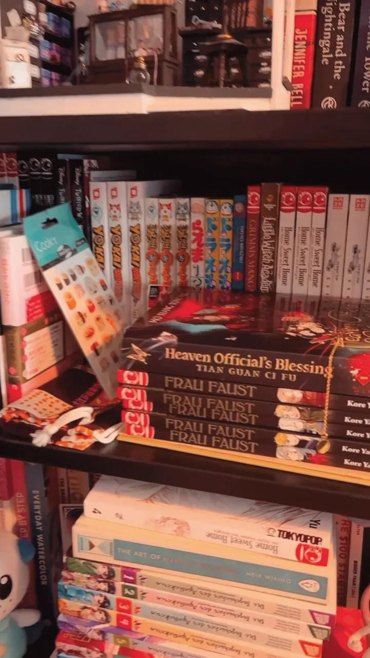 Bücherregal mit Comics, Mangas und anderen Büchern, inklusive 'Heaven Official's Blessing' und 'Frau Faust', sowie einigen Sammelartikeln und Dekorationen.