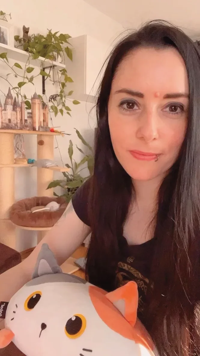 Eine Frau mit langen dunklen Haaren und Piercing im Mund porträtiert sich mit einem lustigen Katzen-Kuscheltier in einem Wohnzimmer.