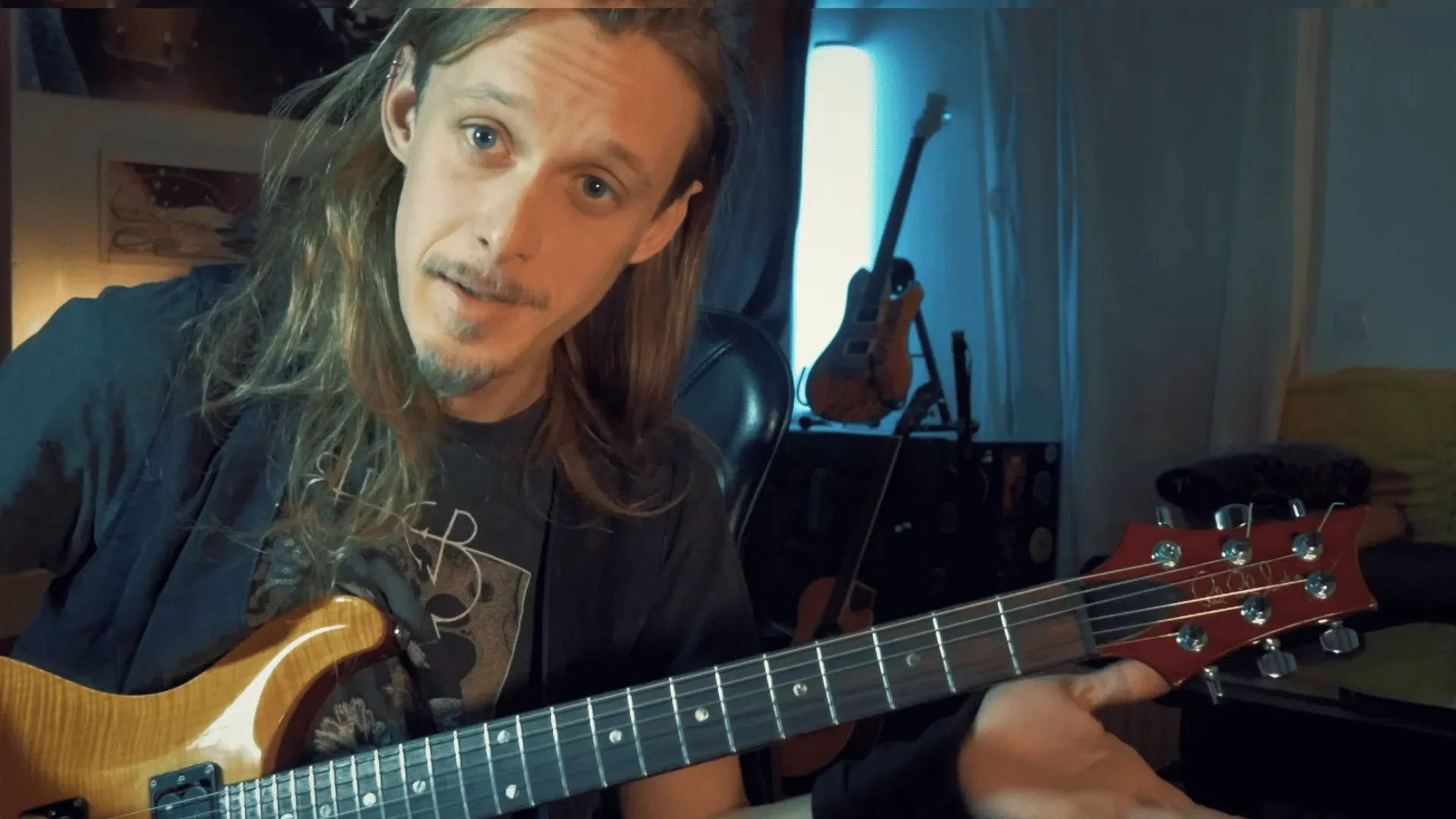 Ein Mann mit langen Haaren und Schnurrbart, der eine Gitarre spielt, in einem Raum mit Musikinstrumenten im Hintergrund.