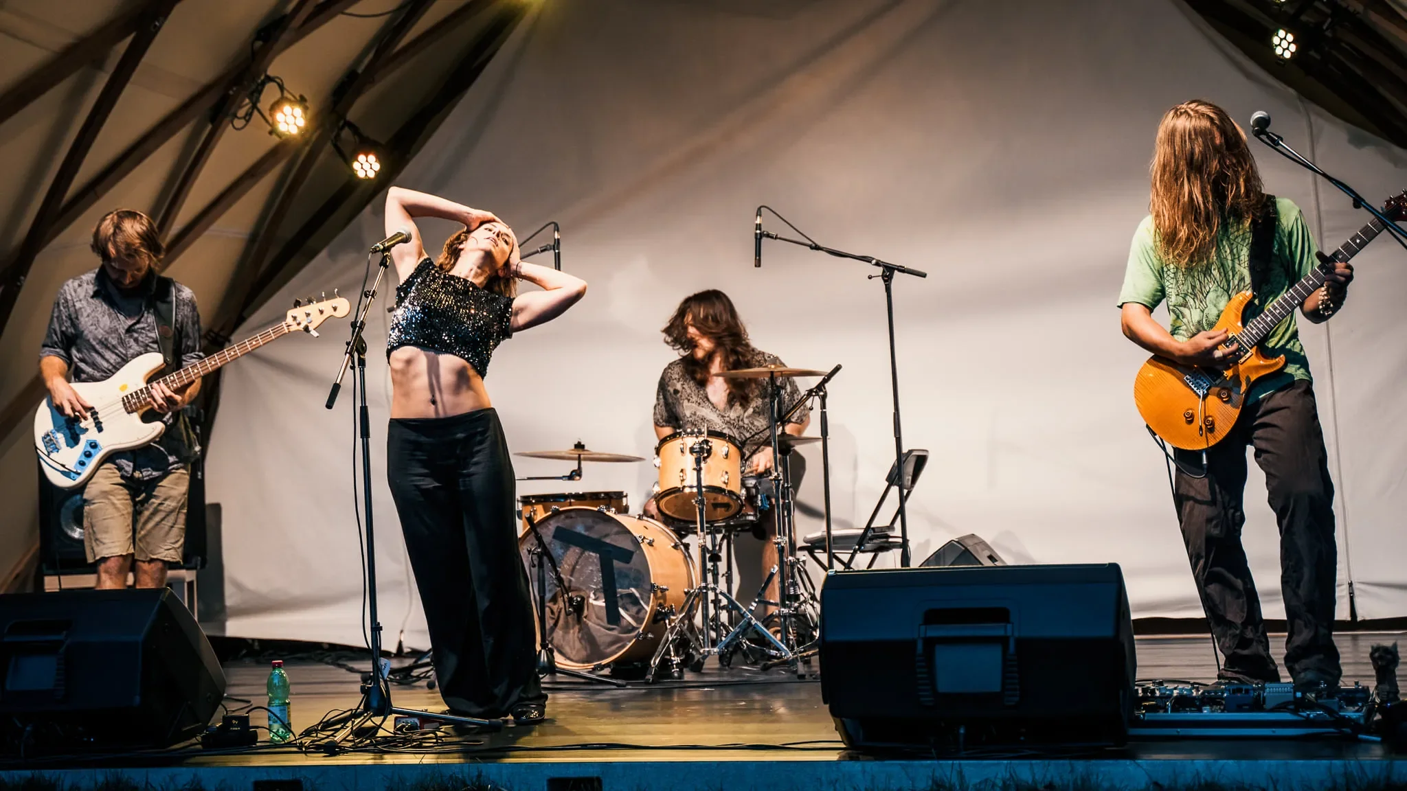 Eine Band mit vier Mitgliedern spielt auf einer Bühne. Ein Mann auf der linken Seite spielt E-Gitarre, eine Frau in der Mitte singt, im Hintergrund spielt ein weiterer Mann Schlagzeug, und auf der rechten Seite spielt ein Mann akustische Gitarre.