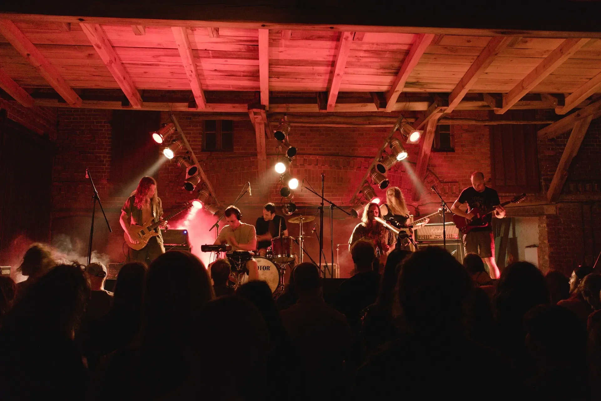Eine live-Band spielt auf einer Bühne in einem Keller mit Backsteinwänden, beleuchtet von rotem Licht, vor einem Publikum.