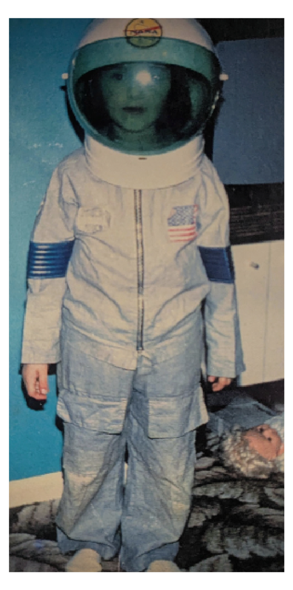 Child dressed in an astronaut costume with a space helmet and American flag patch, standing indoors on a patterned carpet.
