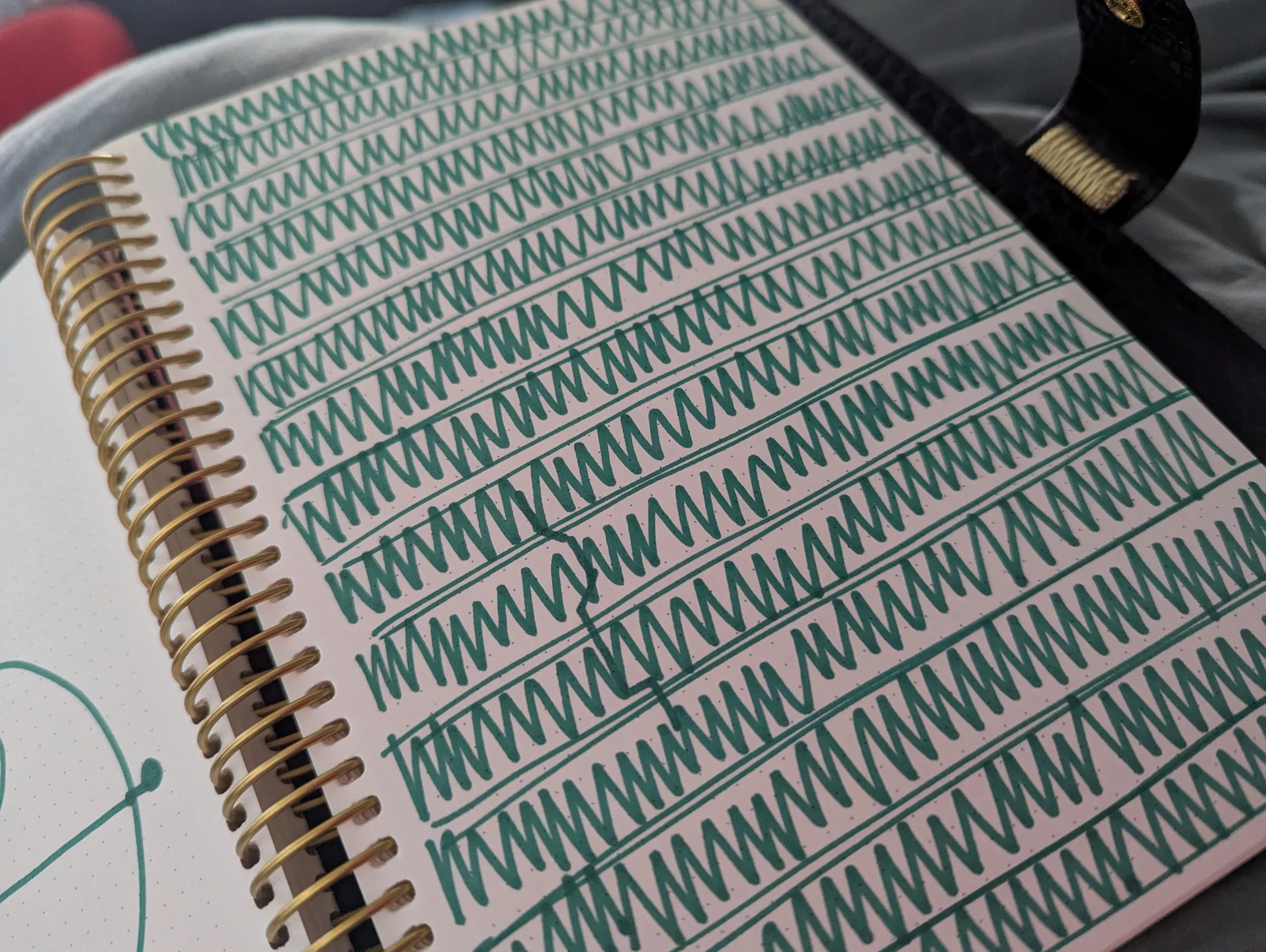Close-up of a notebook page with repeated green doodles of a spiky line pattern, and a black pen with gold accents resting on the page.