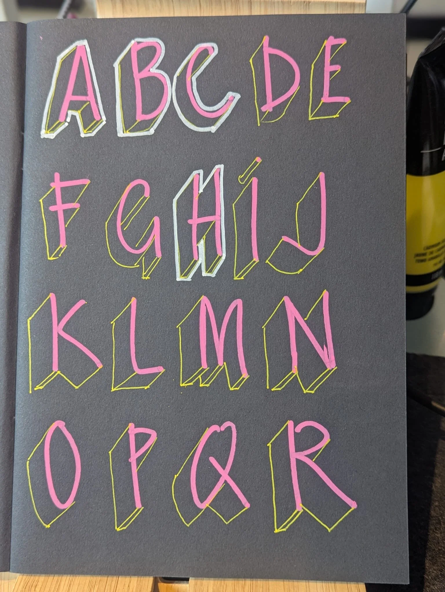 Sign with the alphabet written in three-dimensional pink letters outlined in white and yellow on a dark background.