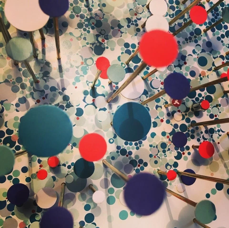 Colorful abstract art installation with various-sized circular discs in shades of blue, red, white, and green, mounted on thin metallic rods in a scattered pattern, creating a layered 3D effect.