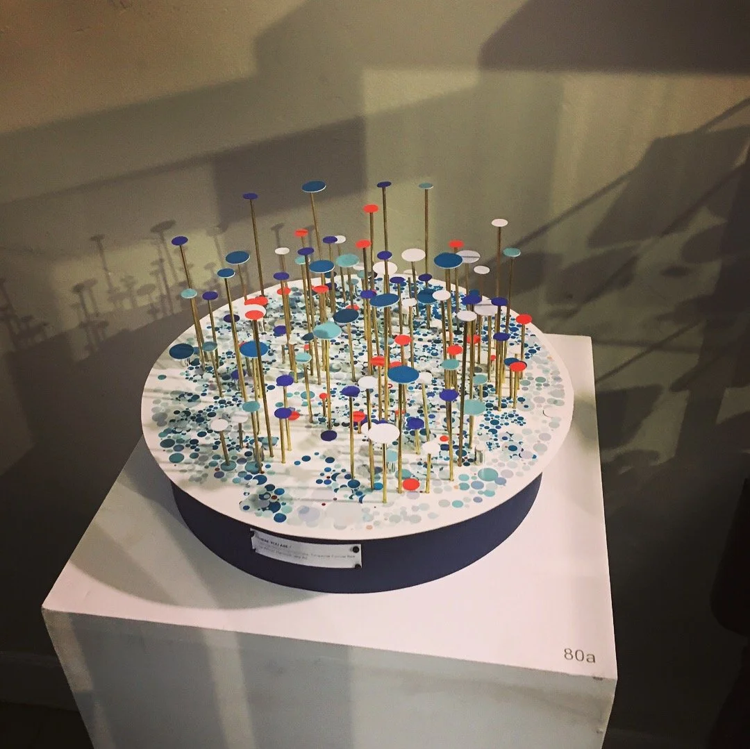 A circular art piece featuring numerous small colored circular elements on slender brass rods, arranged on a surface adorned with small colored dots, displayed on a pedestal in a gallery setting.