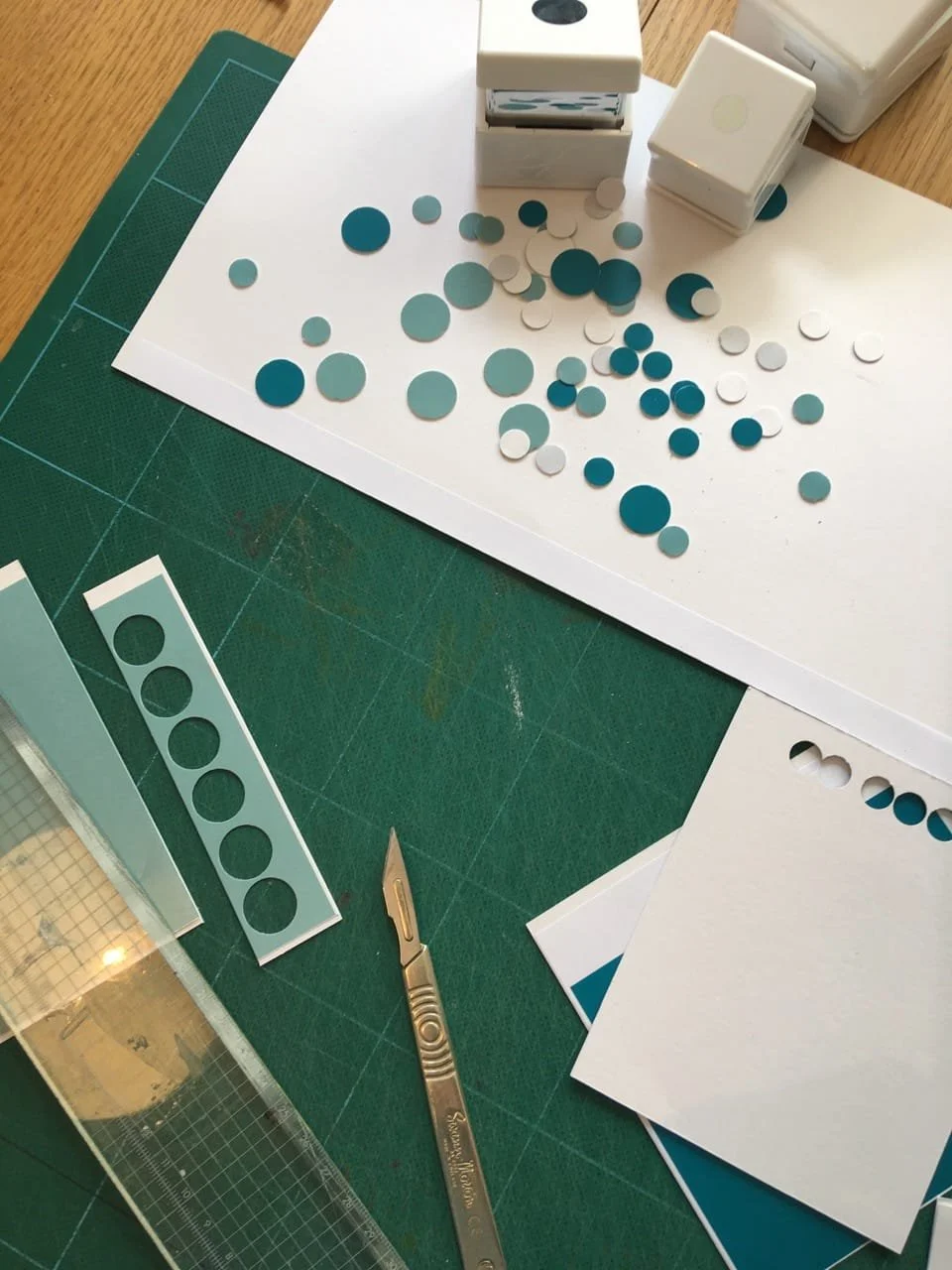 Crafting supplies including white paper with blue and white circular stickers, a precision craft knife, a cutting mat, and sticker sheets, arranged for a project.