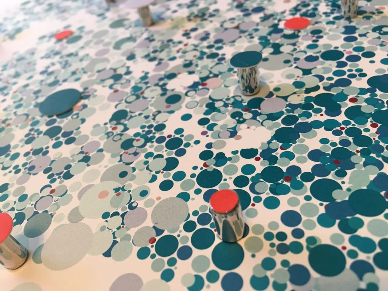 Close-up of a tabletop covered with numerous blue, teal, white, and red circular confetti. There are a few metallic game pieces, one with a red top, another with a blue top, and a taller one with a reflective silver surface. The scene suggests an ong