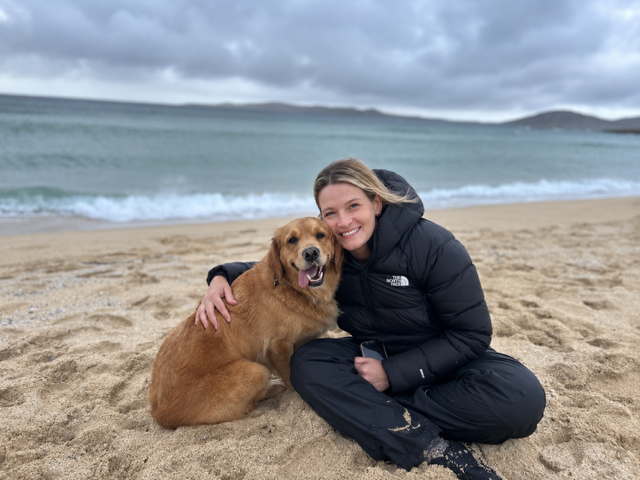 A woman smiling and sitting on a sandy beach next to her golden retriever dog on a cloudy day, near the ocean