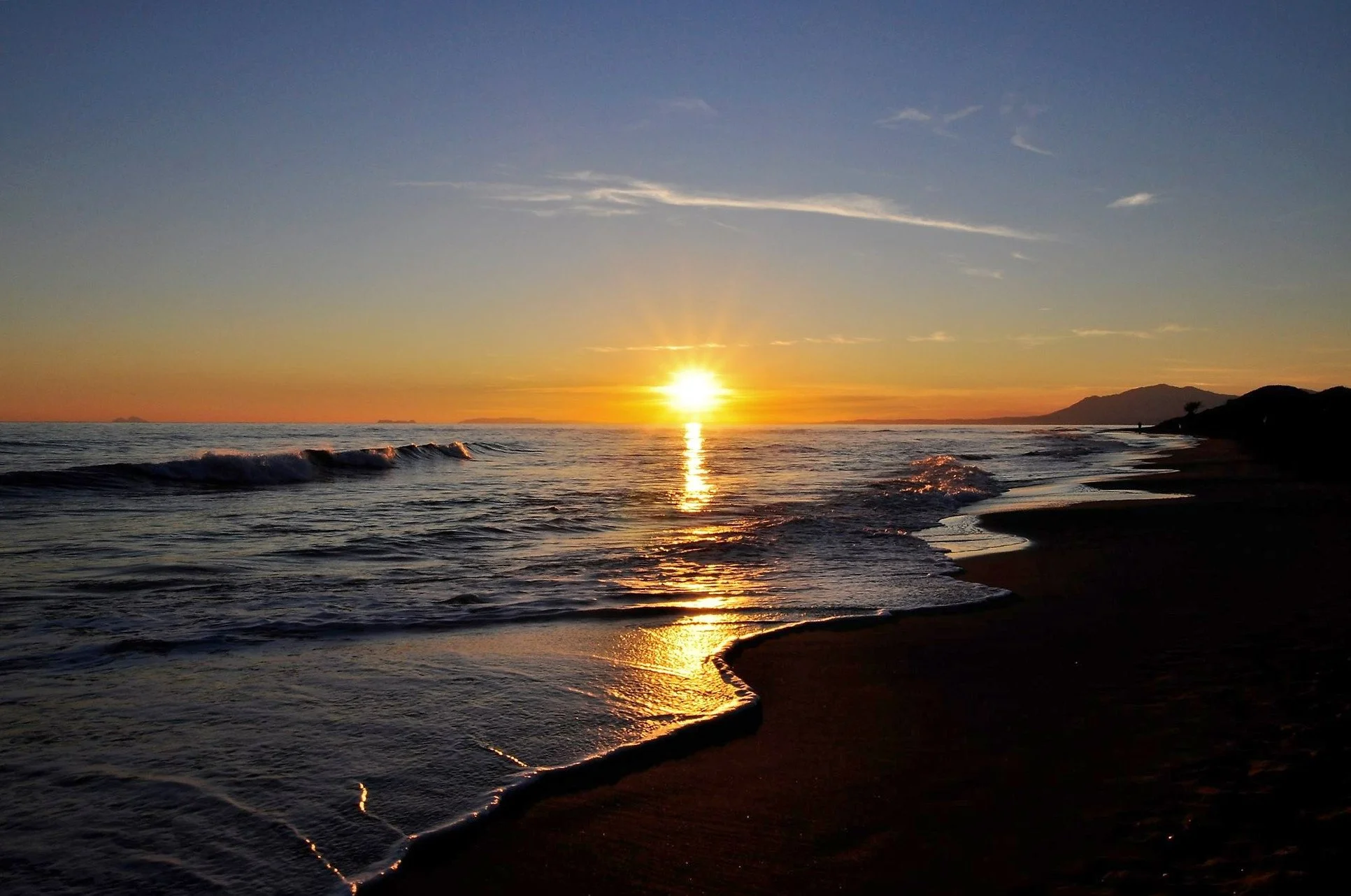 Sunset over the ocean with waves gently crashing on the shore and a silhouette of a mountain in the distance.