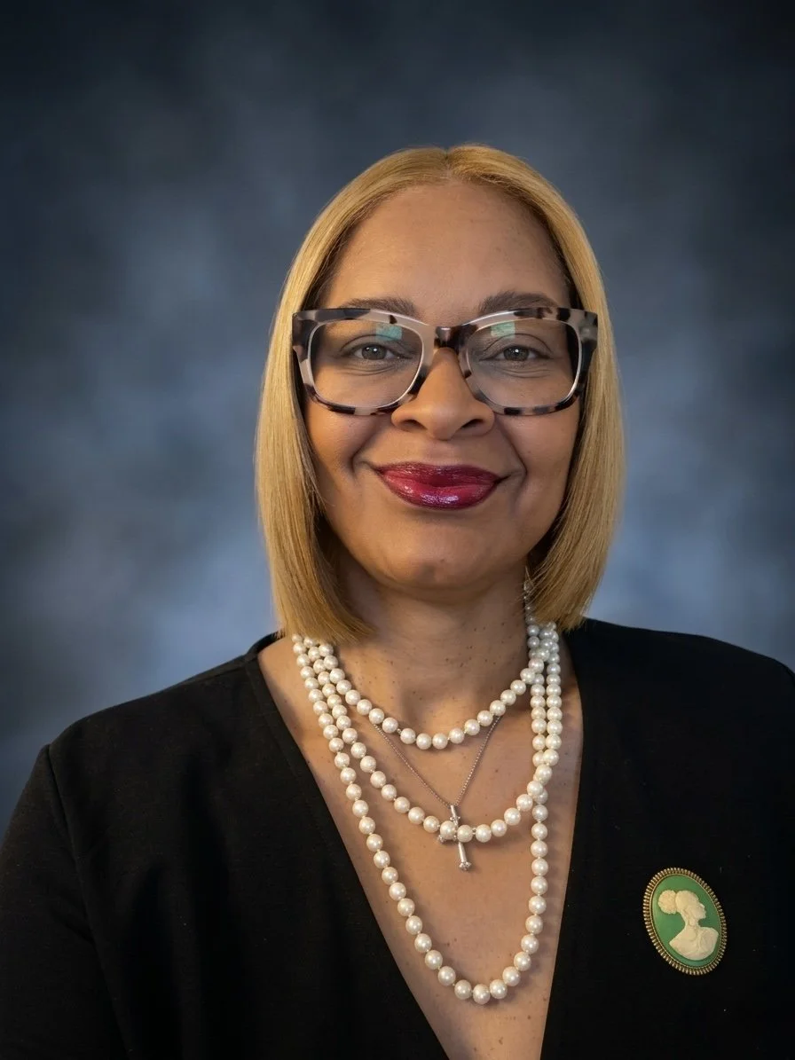 Portrait of a smiling woman with shoulder-length blonde hair wearing large tortoiseshell glasses, red lipstick, multiple pearl necklaces, a black top, and a green brooch with a silhouette of a woman. The background is a blurred dark blue.