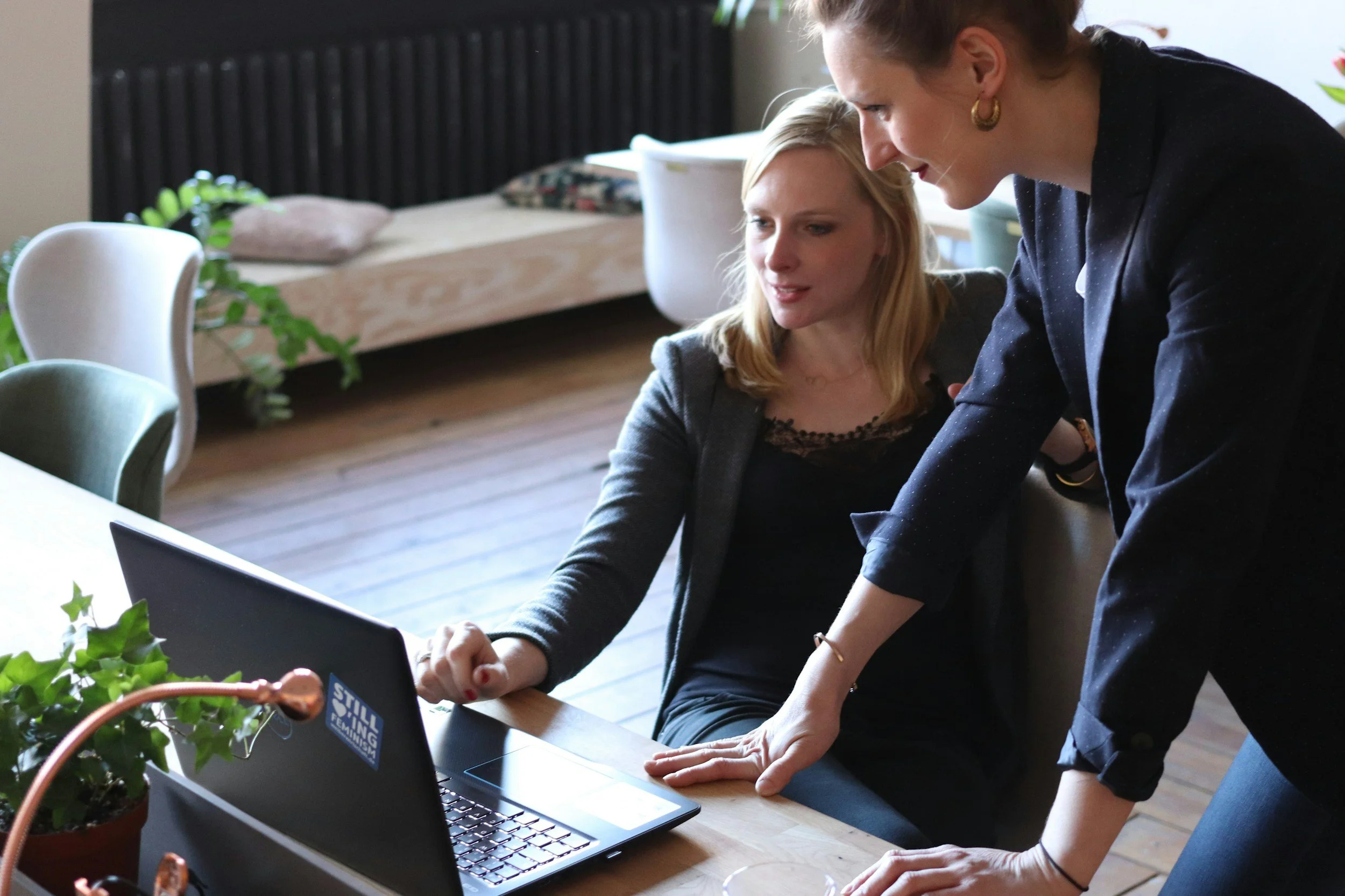Two women working together at a table, looking at a laptop screen, with one woman standing and the other sitting, in a well-lit room with a bed, plants, and chairs in the background.