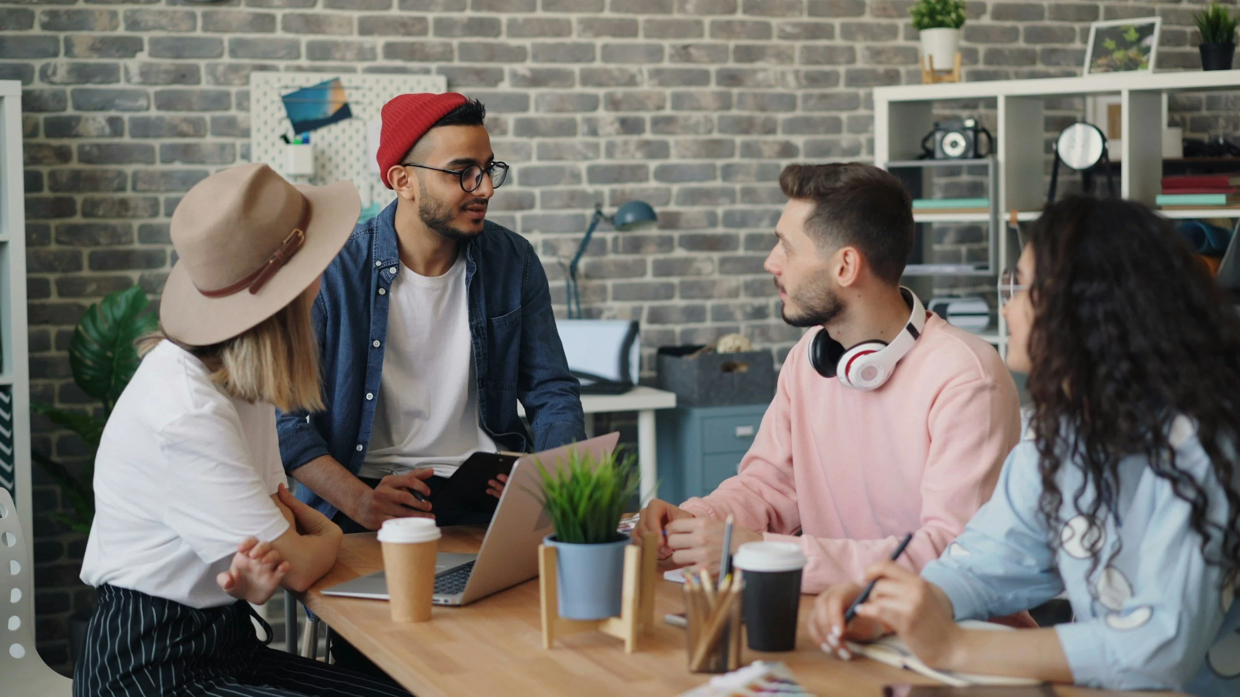 Group of five people having a discussion in a modern office with a brick wall background, plants, and shelves with decorations.