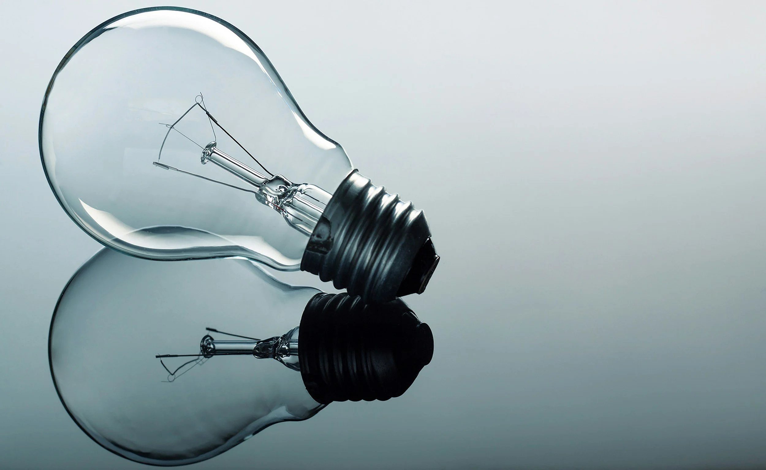 A clear incandescent light bulb lying on a reflective surface with a plain gray background.