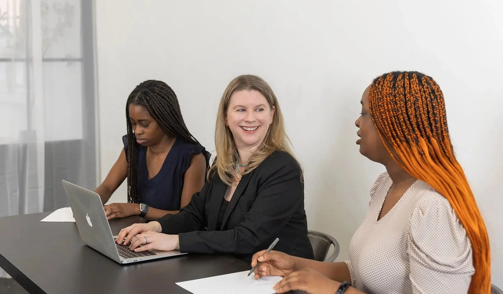Three women sitting at a conference table, two of them taking notes, with one smiling and looking at the other, in a professional setting.
