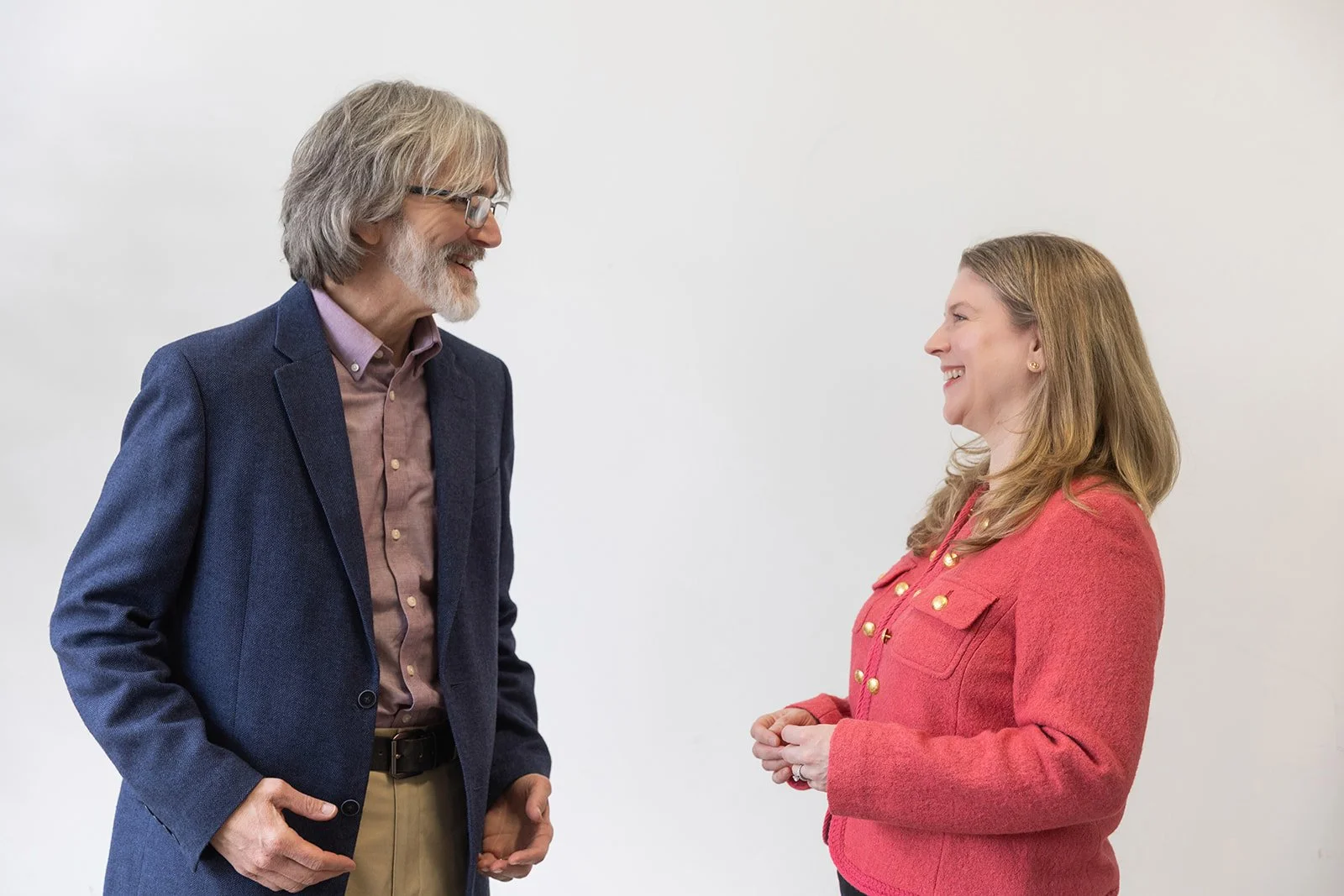 A middle-aged man with gray hair and beard wearing glasses, a navy blazer, and khaki pants, smiling while talking to a woman with long, wavy blonde hair wearing a coral-colored blazer, also smiling, against a plain white wall.