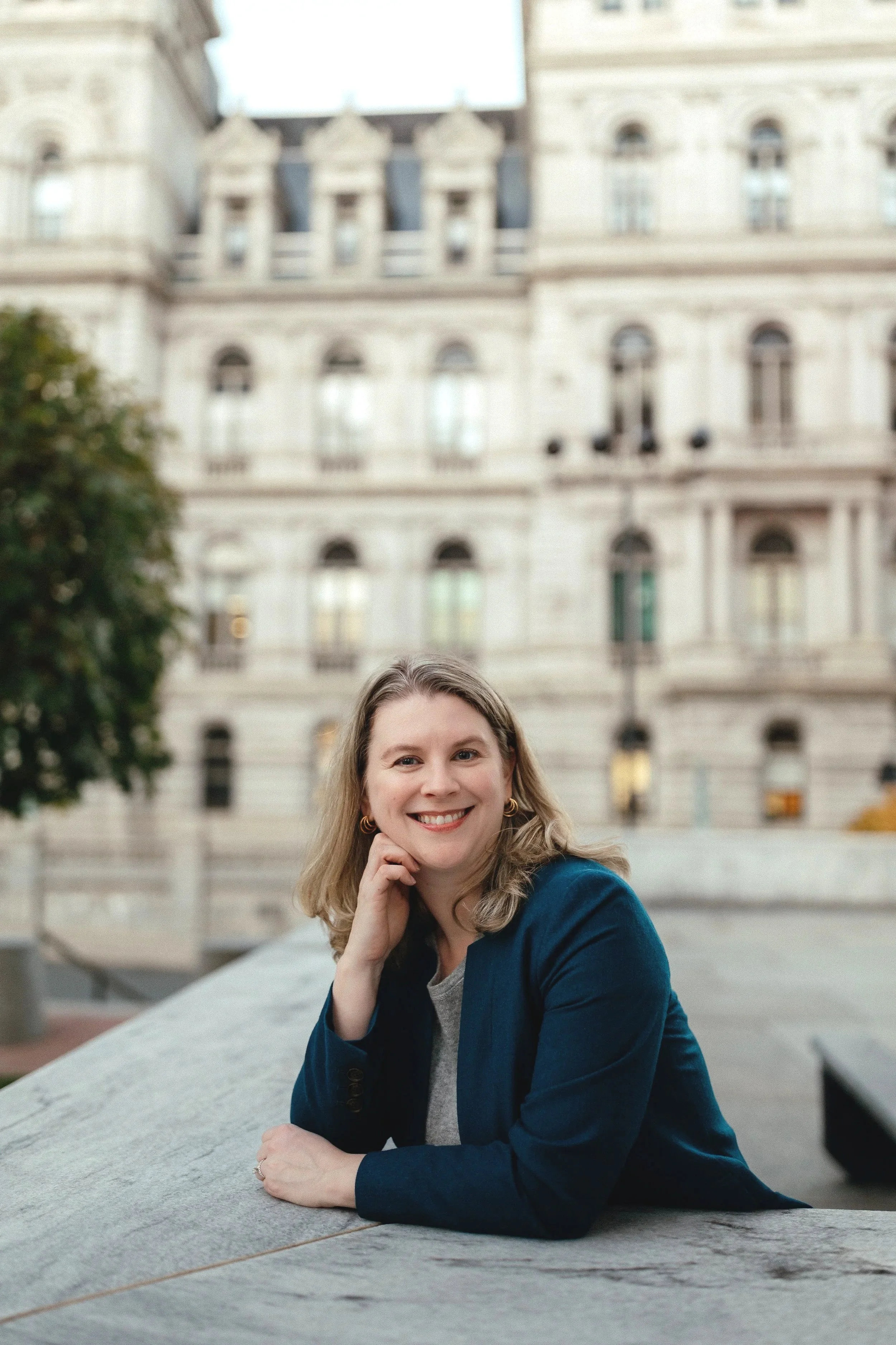 A woman with shoulder-length blonde hair, wearing a blue blazer, smiling and resting her chin on her hand, sitting outdoors in front of a historic building with multiple arched windows.