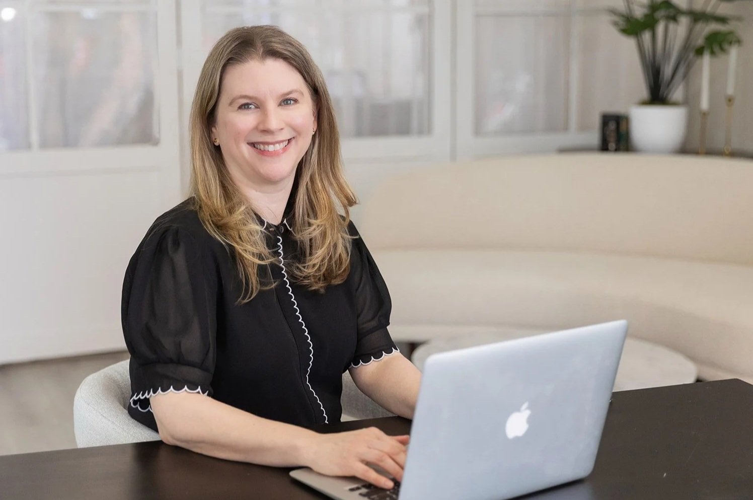 A woman with long blond hair smiling while sitting at a desk with a silver MacBook laptop, in a bright room with large windows and white furniture.