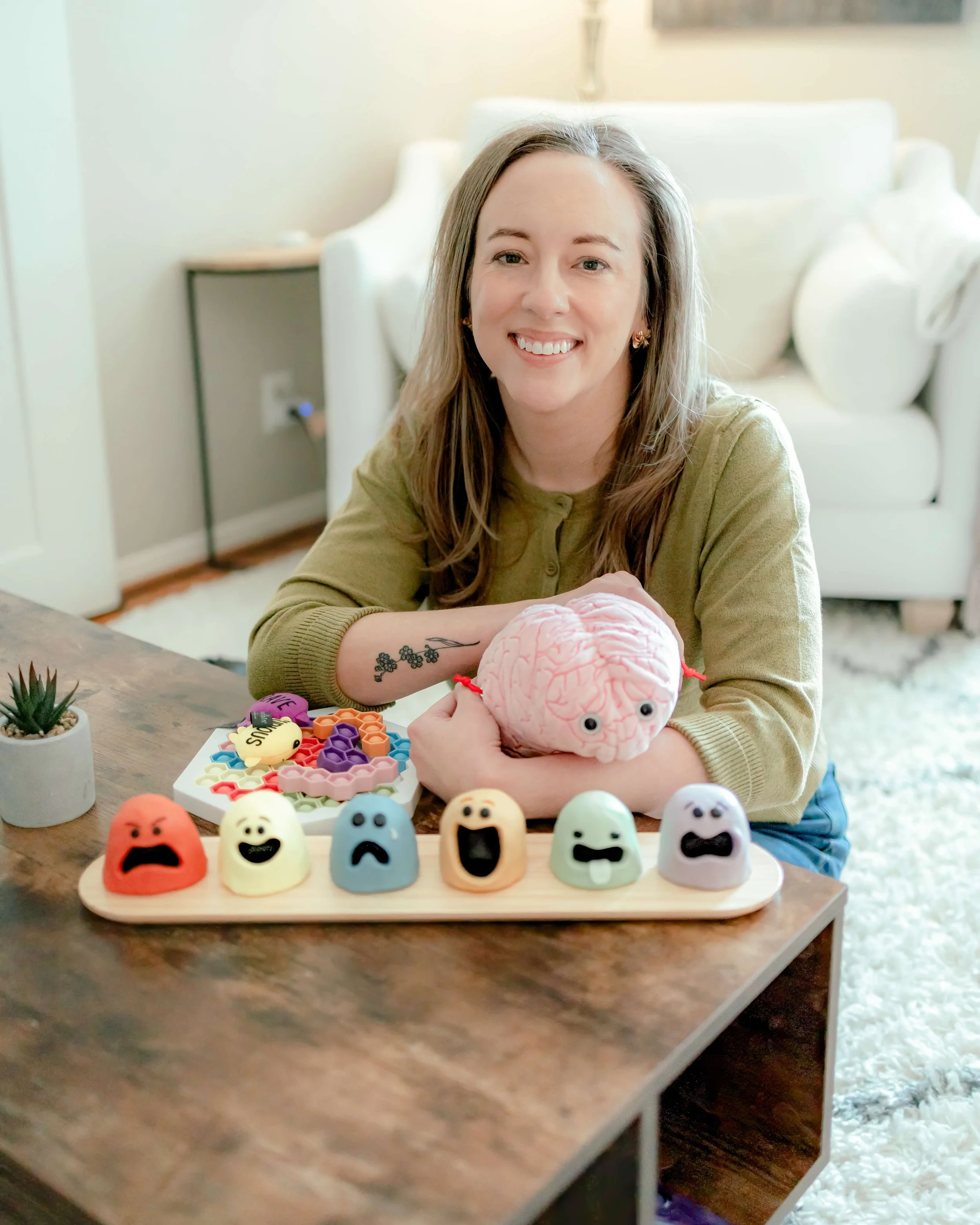 A smiling woman sits at a table with a variety of colorful stress toys and a plush brain on display. The toys are shaped like faces expressing different emotions. In the background, there is a couch and some decorative items.