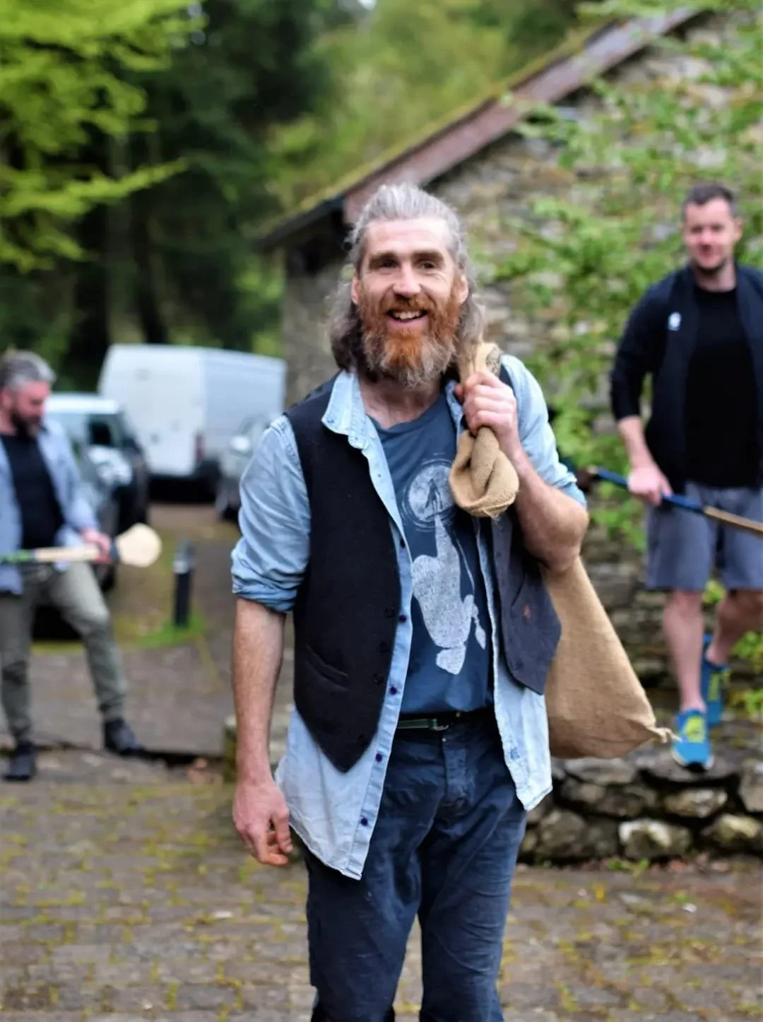 A group of men outdoors with hurling sticks, with two men in the background smiling; additional man in foreground is smiling and has long hair and beard.