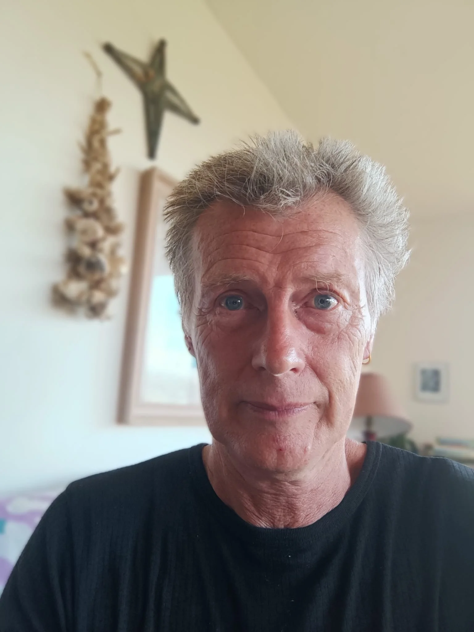 Close-up of a man's face, showing blue eyes and gray hair, wearing a black jumper in a room with framed art and a Brigid's Cross on the wall.