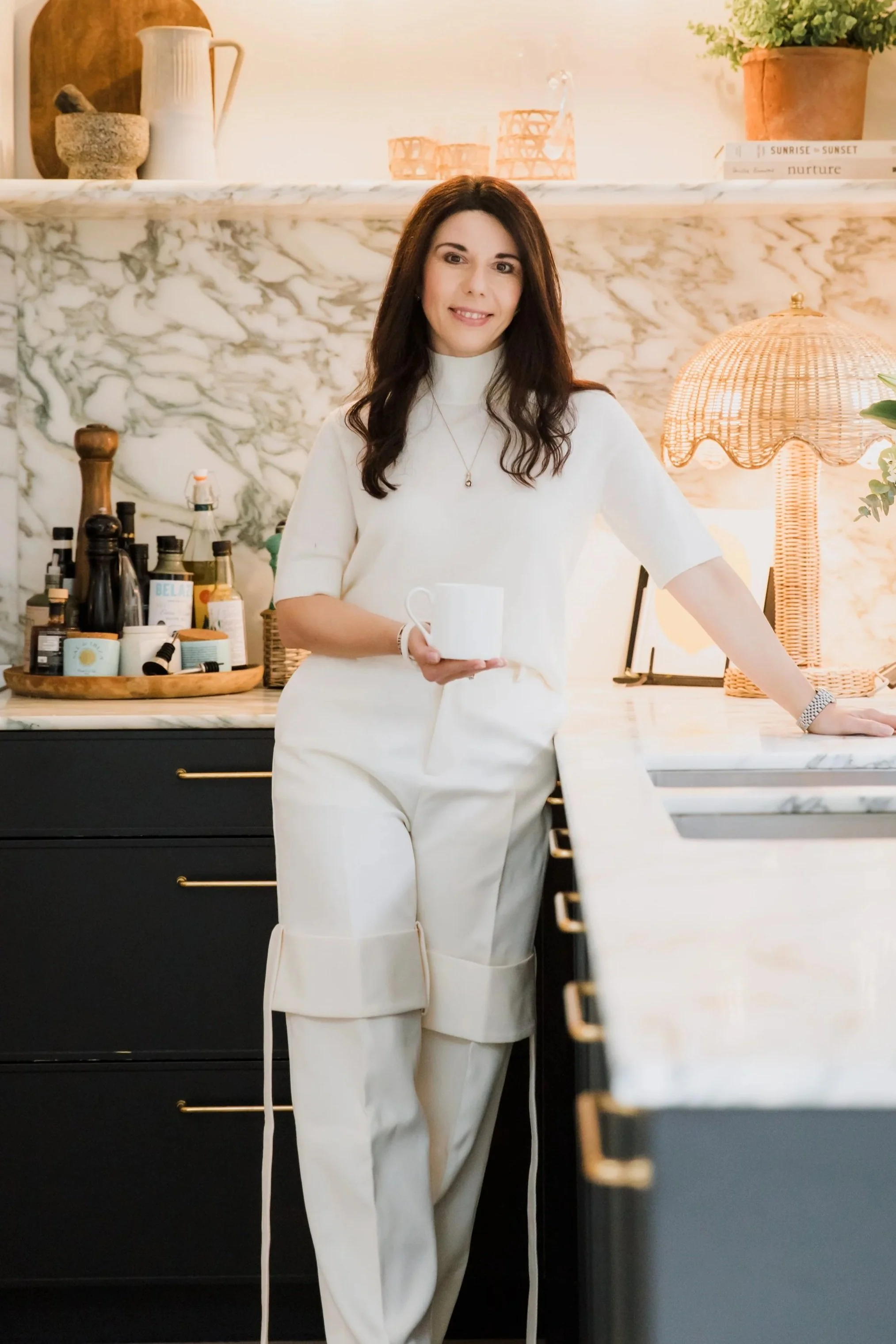 A woman in a white outfit standing in a kitchen with a marble wall, holding a white mug, surrounded by kitchen items and a wicker lamp.