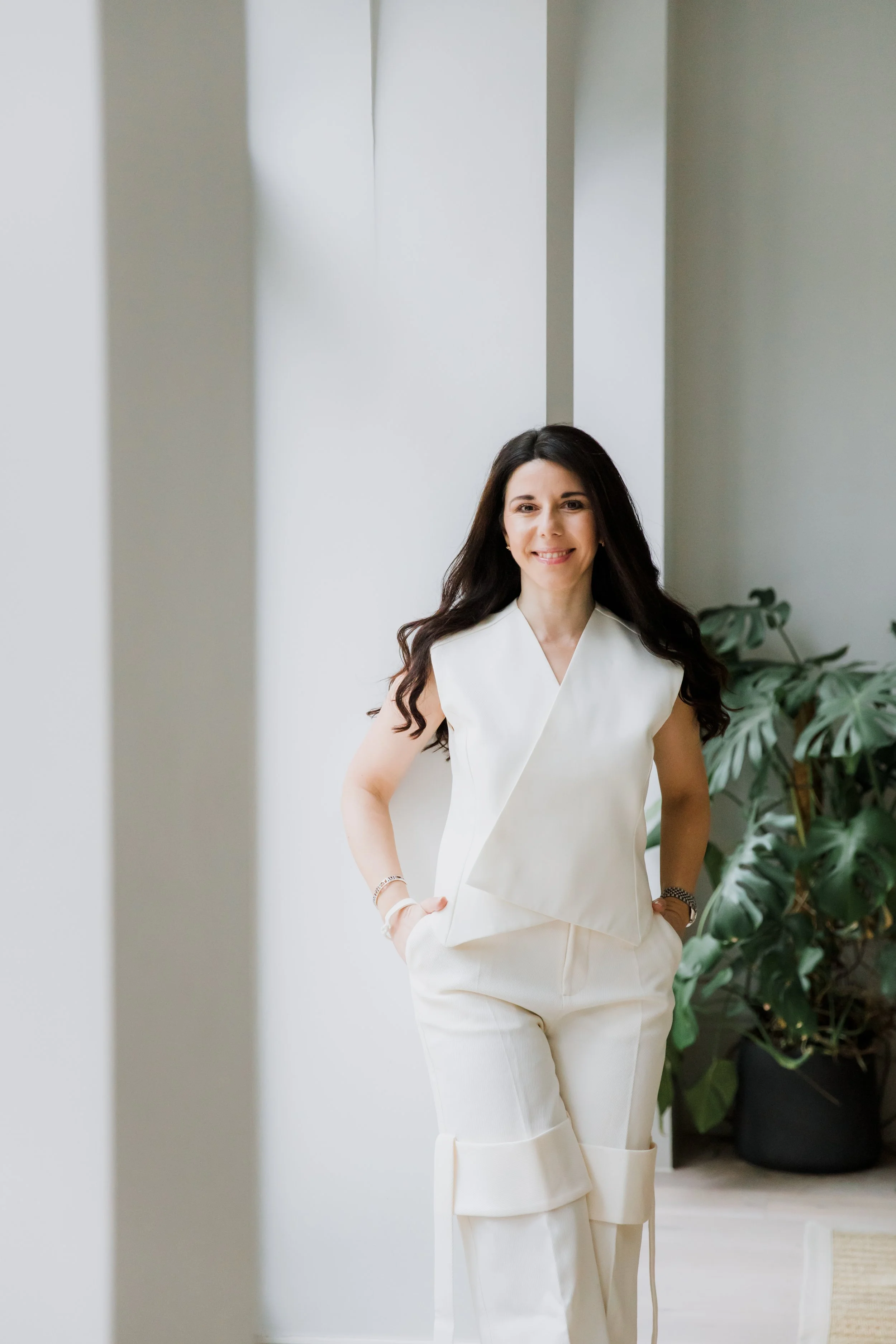 Woman with long dark hair smiling, wearing an elegant white sleeveless top and cream pants, standing indoors near large plant, in a well-lit modern space.