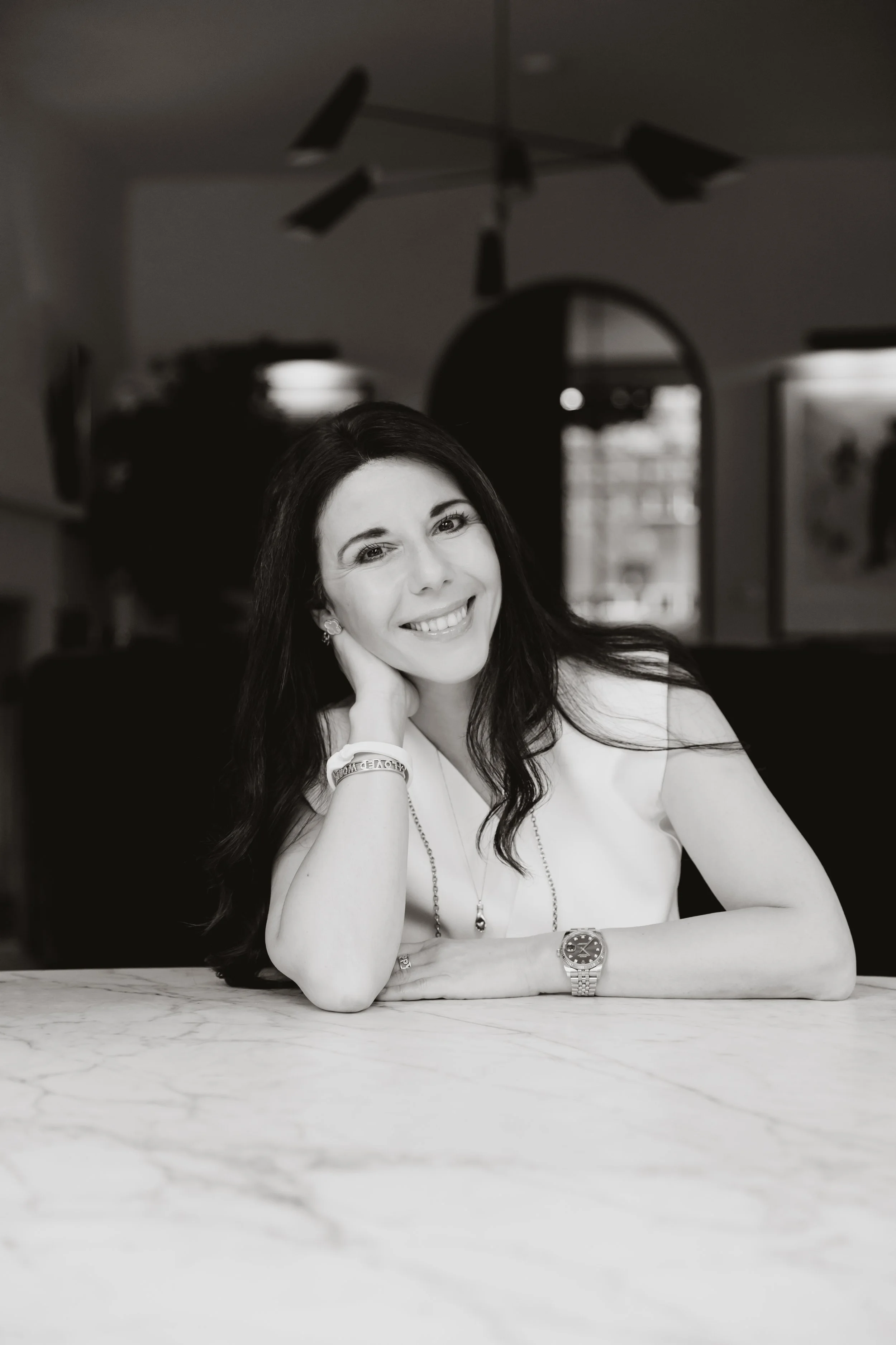 Black and white photo of a woman with long dark hair, smiling, resting her head on her hand, sitting at a table with a marble surface. She is wearing a white top, a wristwatch, a bracelet, earrings, and a necklace. In the background, there is a blurred room with windows and modern ceiling lights.