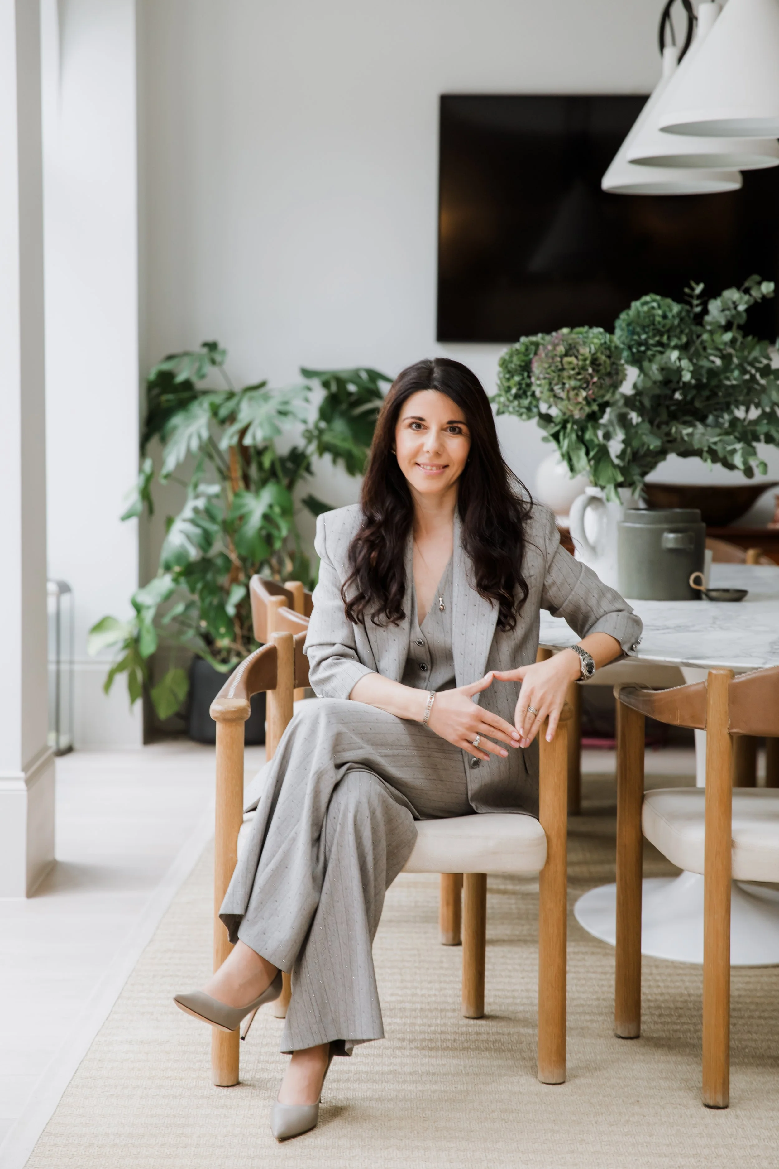 Woman in gray business suit sitting on a dining chair in a modern, well-lit room with green plants and a large black TV on the wall.