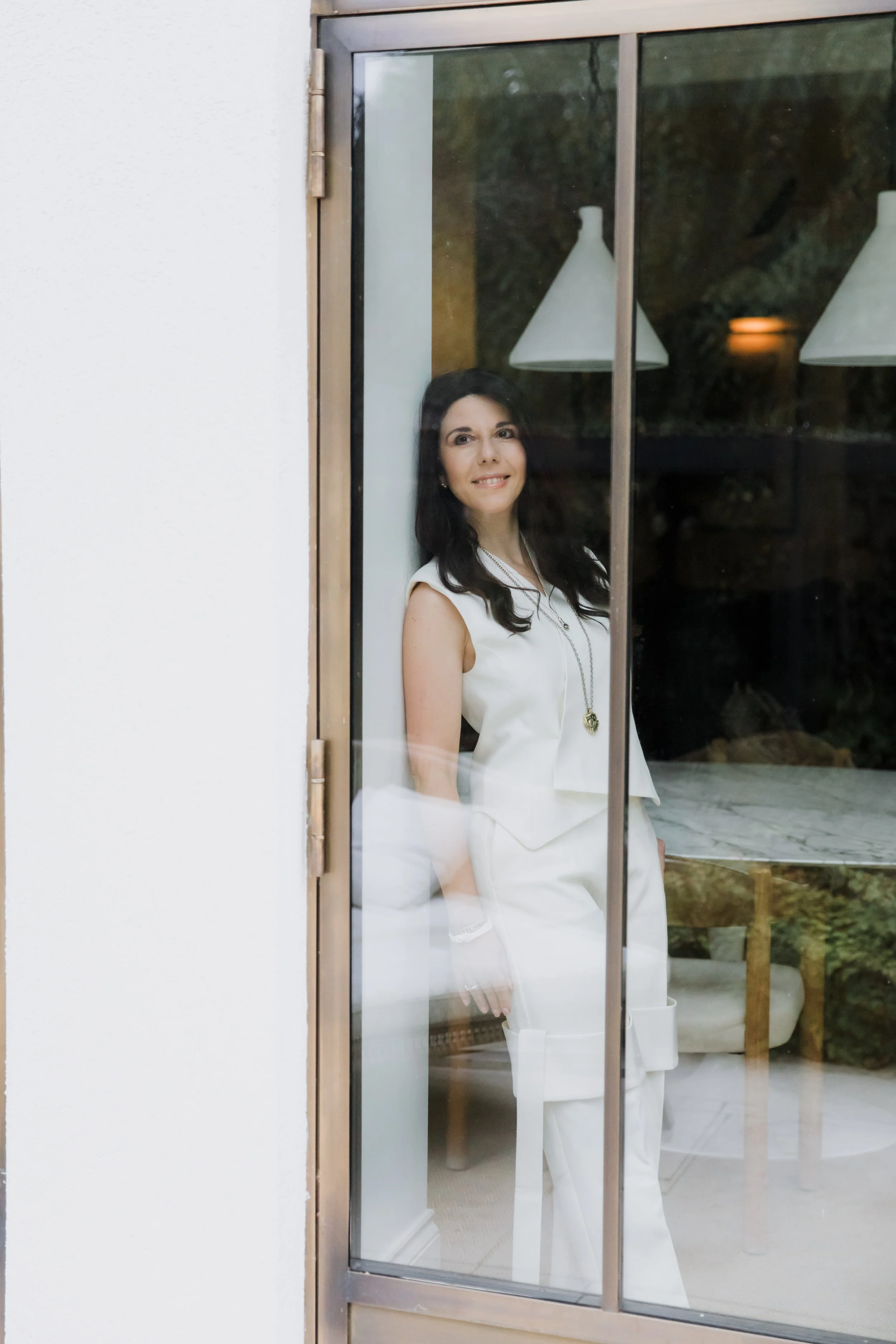 A woman with dark hair and a white sleeveless top standing inside a room, viewed through a glass sliding door, with a background of a bed and hanging lamps.