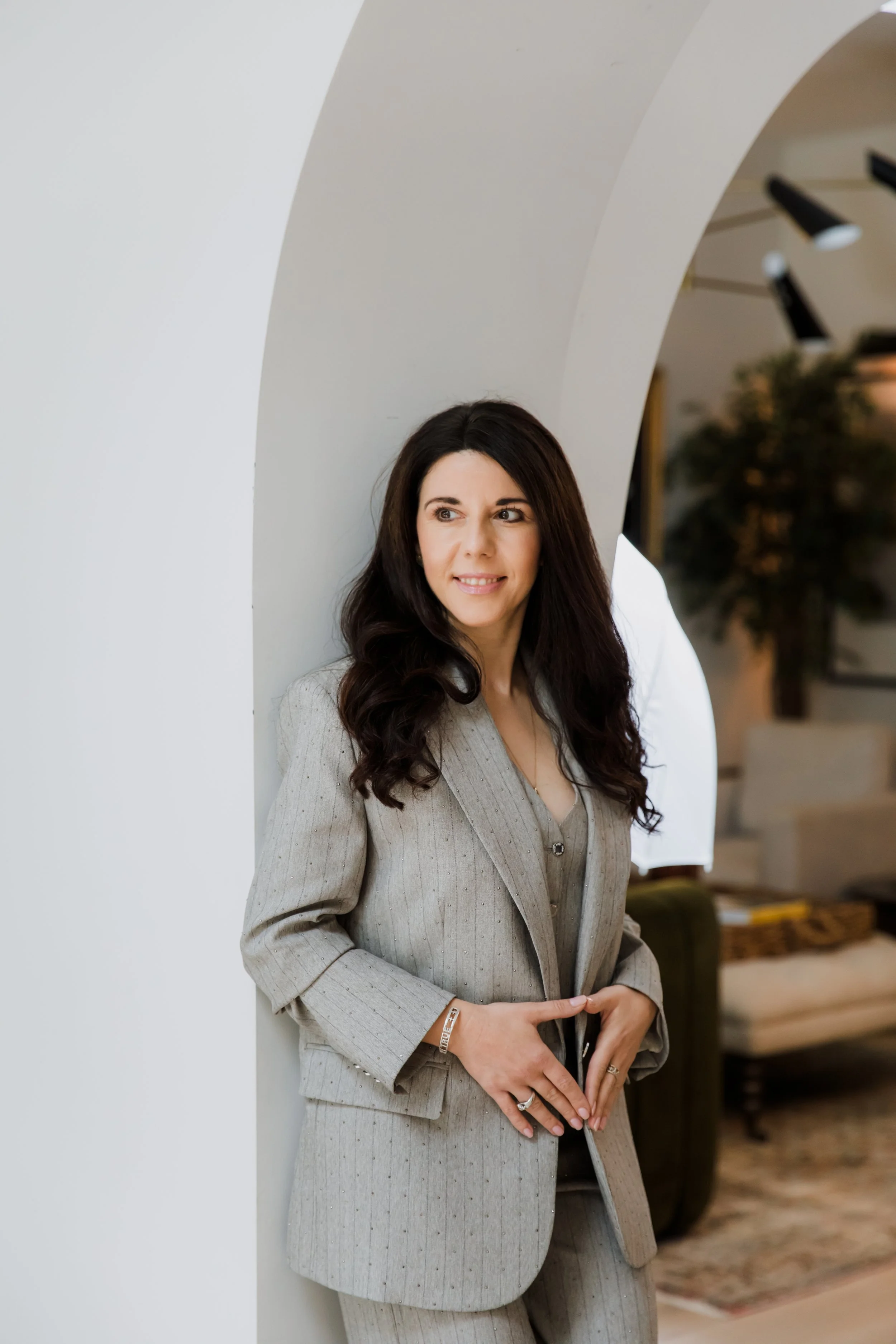 A woman with long dark hair wearing a light gray pinstripe suit, standing in front of a white wall with an arched ceiling, in what appears to be an indoor setting with blurred background.