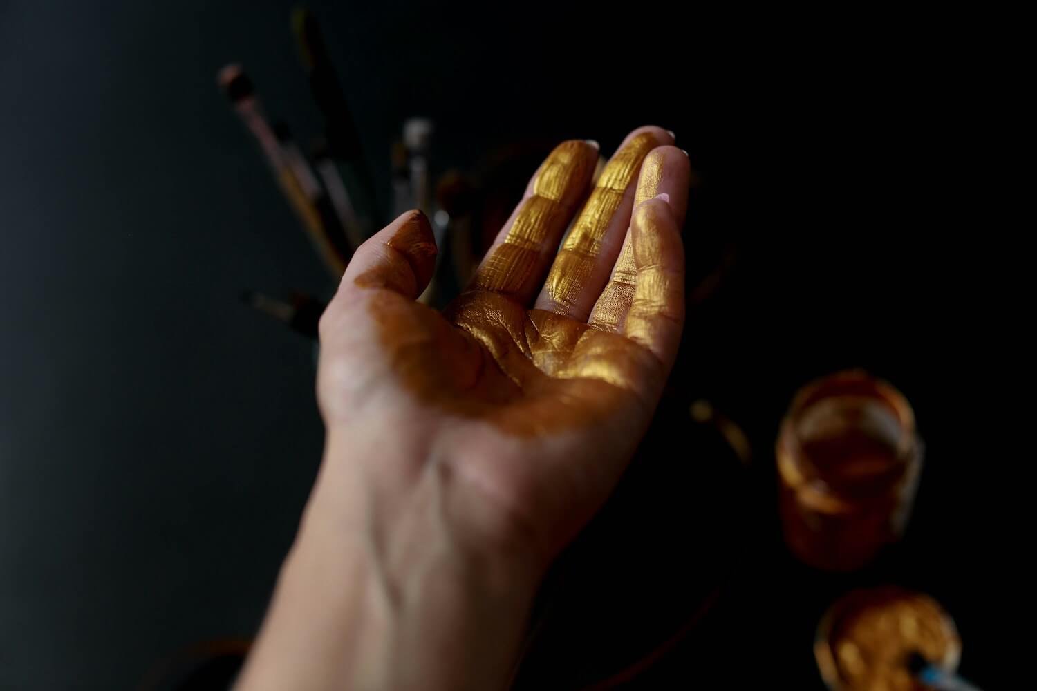 A person's hand covered in gold paint, reaching out over a black surface with art supplies and jars of gold paint nearby.