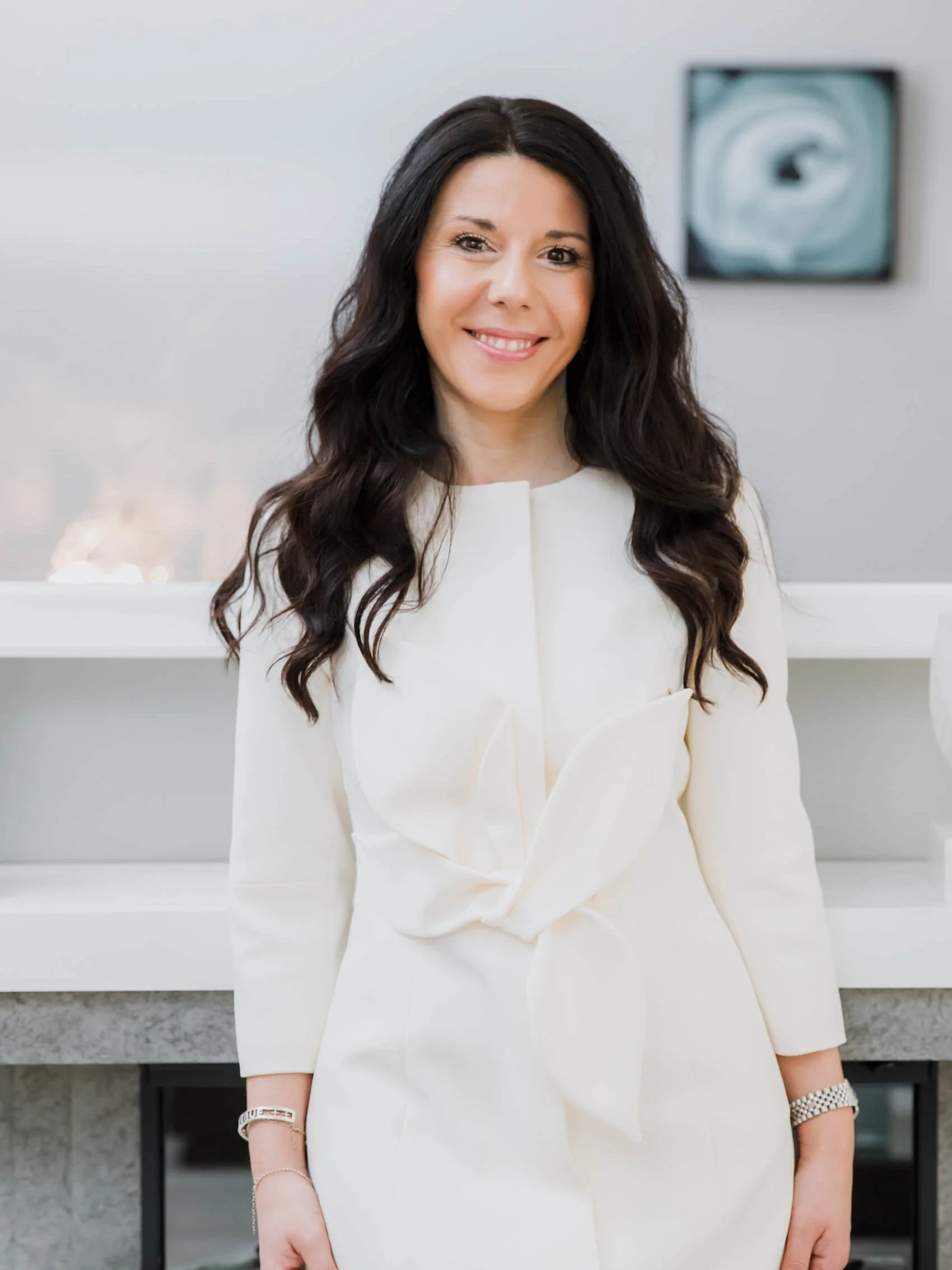 Portrait of a smiling woman with long dark wavy hair wearing a white blazer and accessories, standing in front of a modern office background.