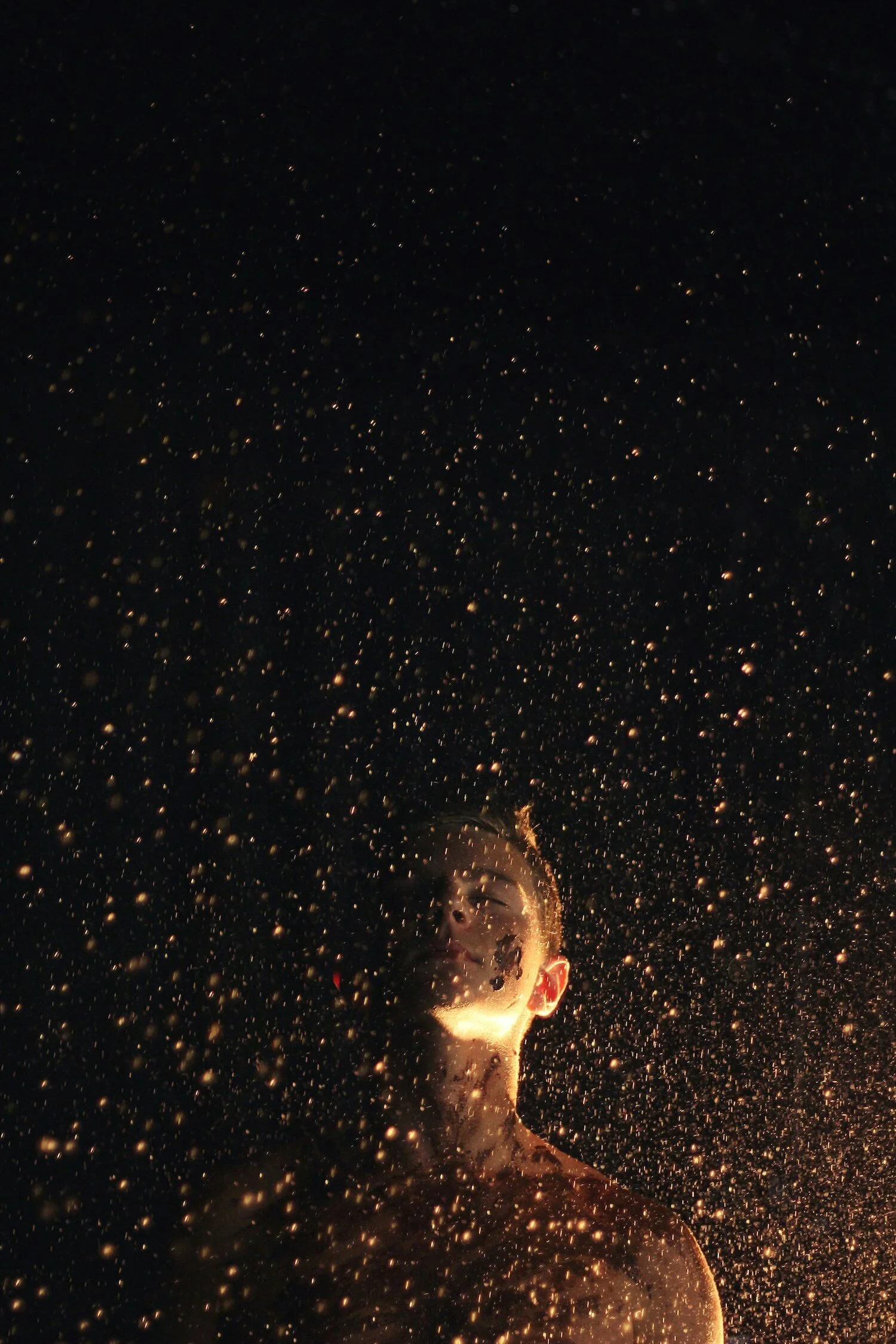 A person with glasses and short hair is illuminated by sparks from a firework, with a shower of sparks surrounding them in a dark background.