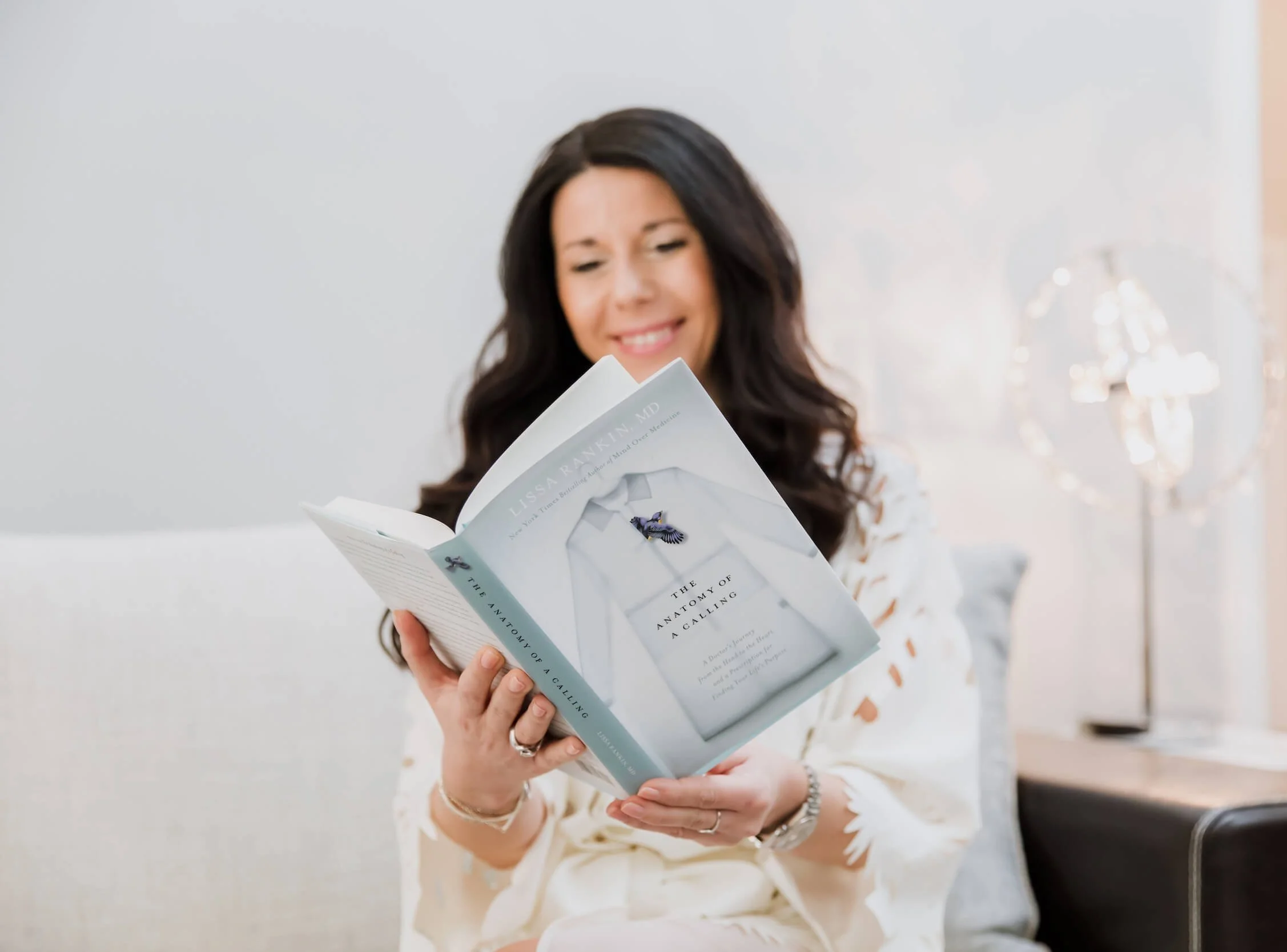 A woman with long dark hair, wearing a white dress with orange spots, is smiling while reading a book titled "The Anatomy of a Calling." She is in a bright room with a white wall and a lamp in the background.
