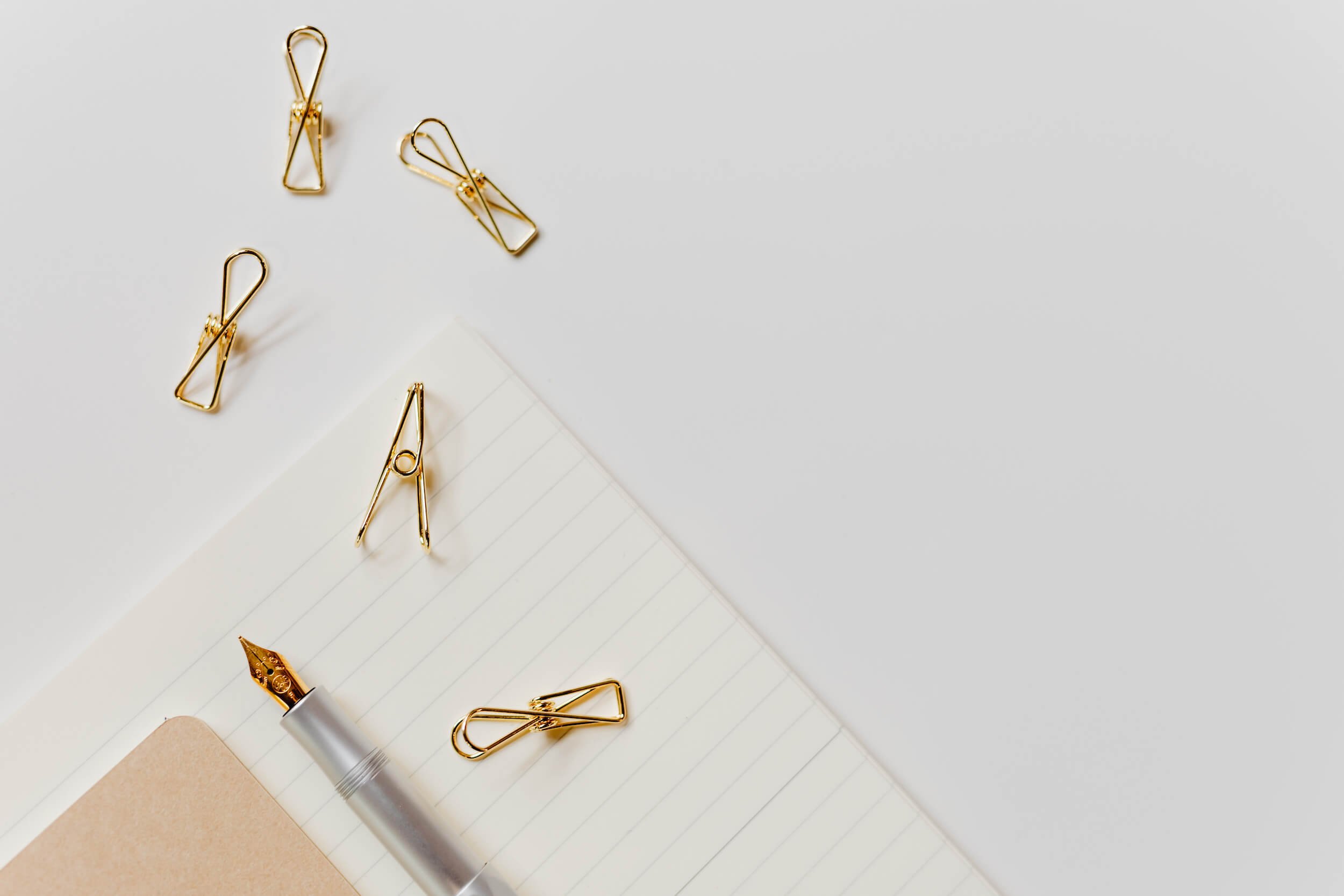 Golden paper clips scattered on lined paper, with a silver and gold pen and a brown piece of paper underneath.