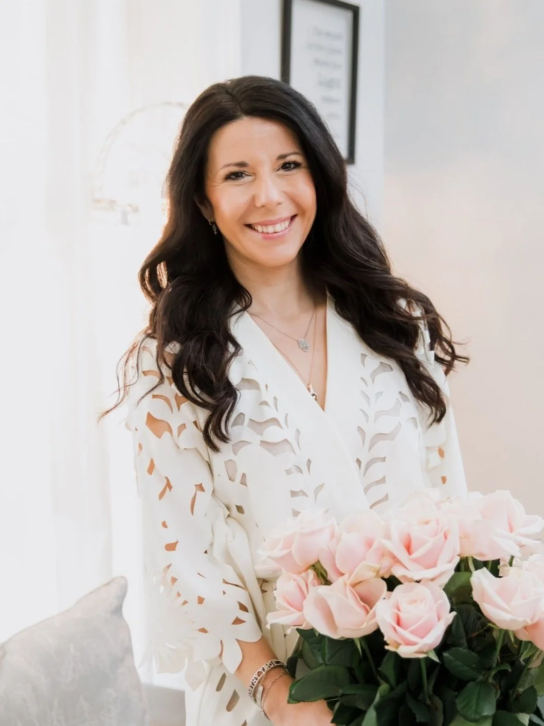 A woman with long dark hair smiling, holding a bouquet of pink roses, in a well-lit room with framed artwork on the wall.