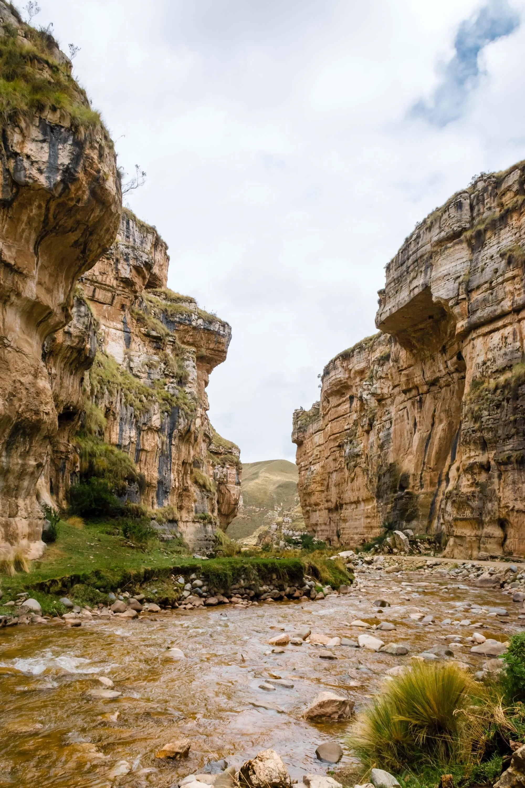 A deep canyon with steep, layered rocky walls on both sides, a small stream flowing at the bottom, and some green vegetation near the stream, under a partly cloudy sky.
