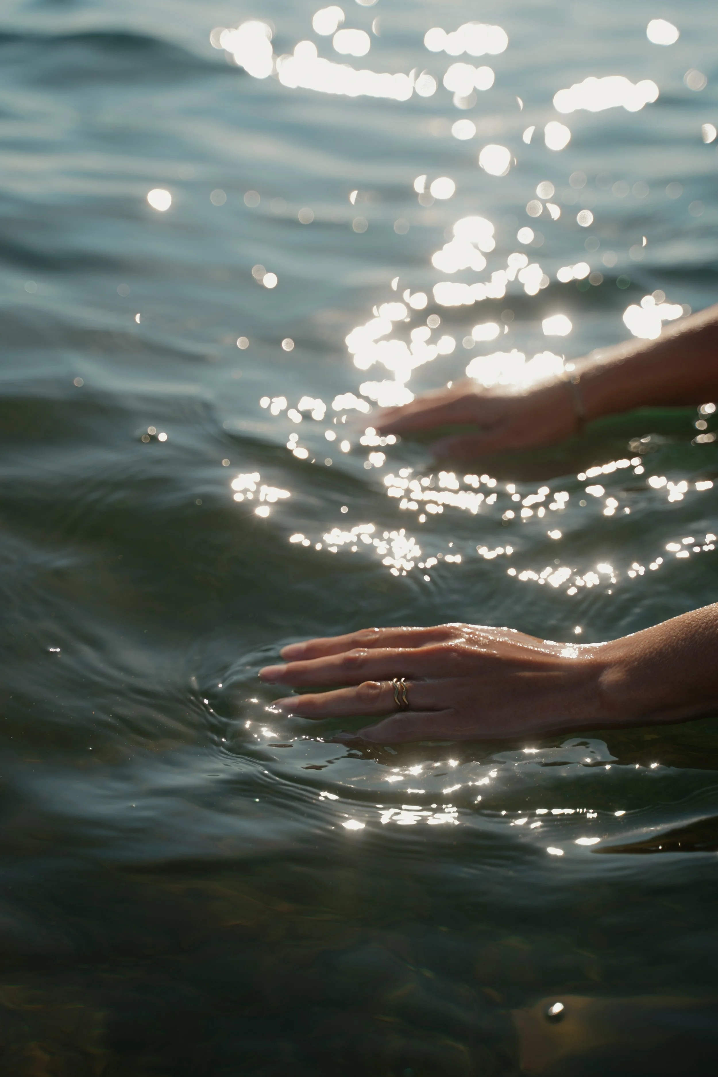 Close-up of a person's hand with rings, floating on water with sunlight reflecting off the surface.