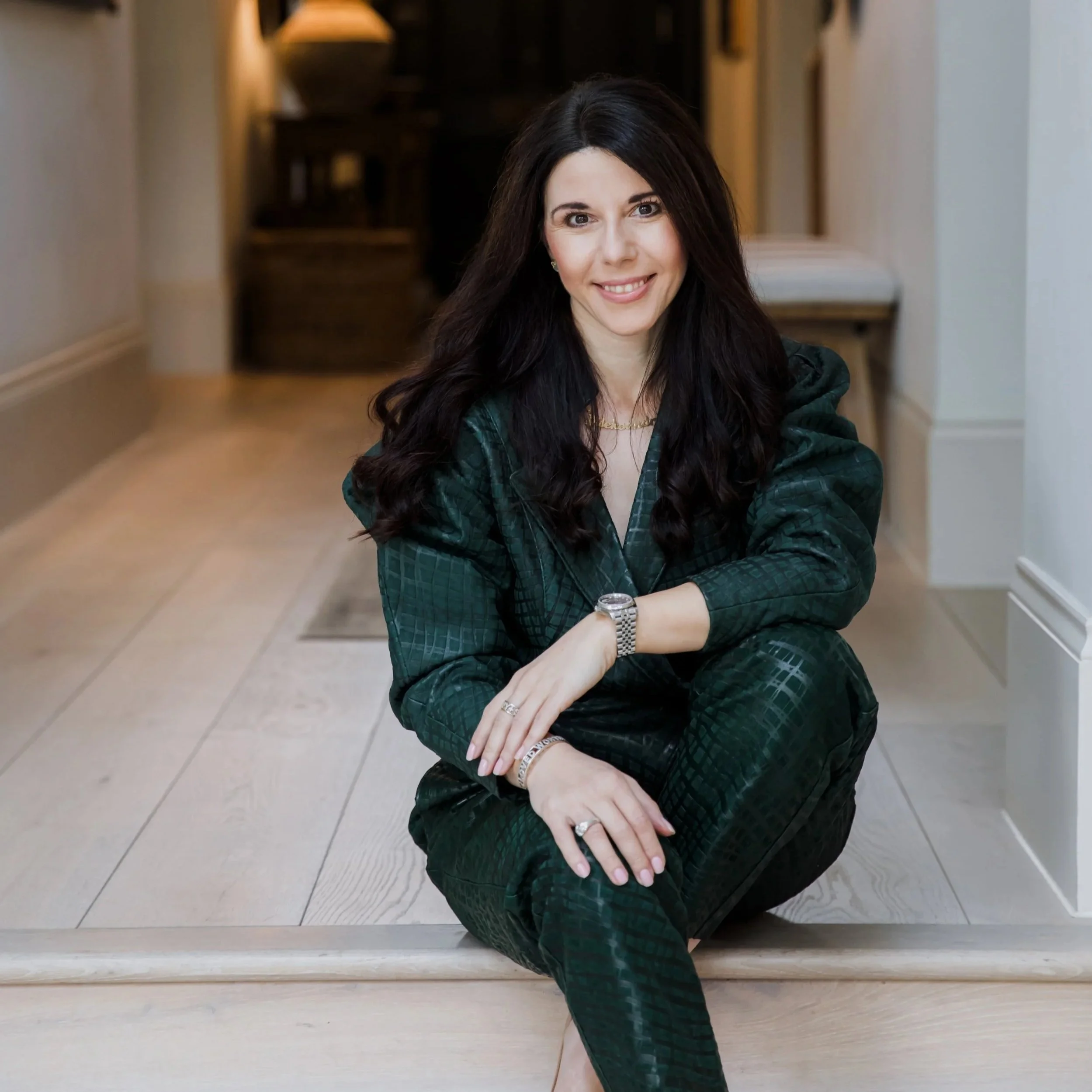 A woman with long dark hair smiling, sitting on the floor in a hallway, wearing a green plaid blazer, with jewelry including a watch and rings, and looking at the camera.