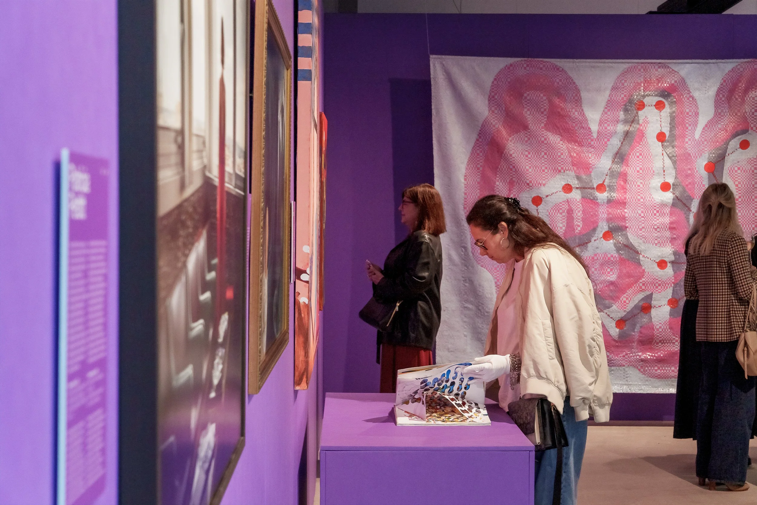 Pessoas olhando quadros em uma galeria de arte com paredes roxas e uma obra com fundo branco e detalhes em rosa, com pontos vermelhos conectados por linhas.