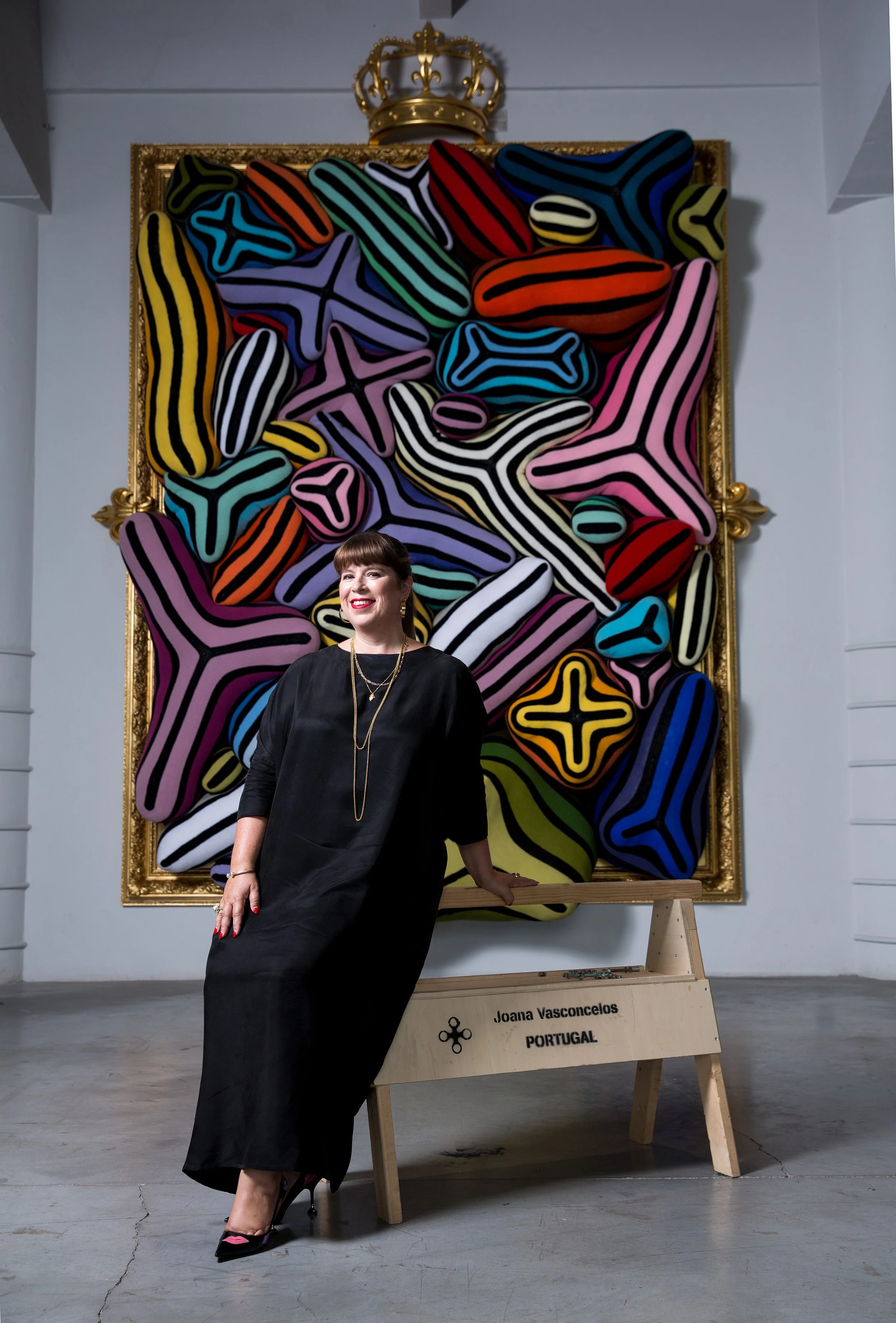 Mulher sorridente com vestido preto e salto alto, posando ao lado de uma escultura feita de formas coloridas e linhas pretas, em uma galeria de arte. Na escultura, há uma placa com o nome Joana Vasconcelos, de Portugal.