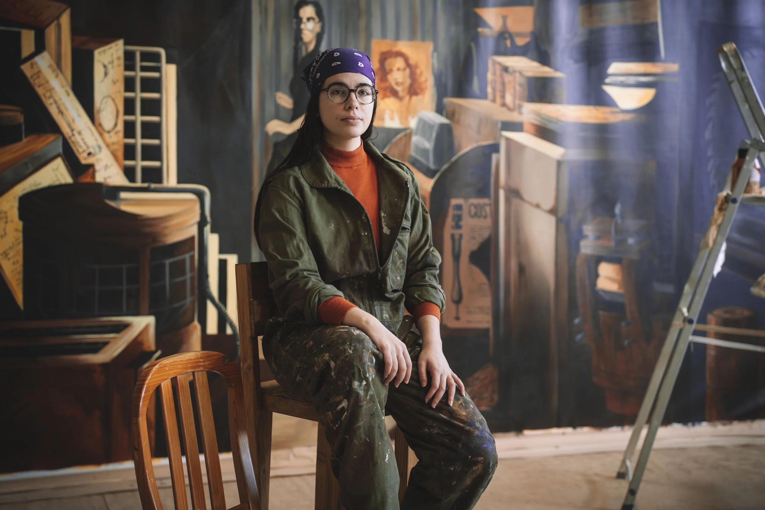 Mulher com bandana roxa, óculos e roupa de camuflagem, sentada em uma cadeira de madeira, em um ambiente com paredes de madeira e obras de arte, incluindo um quadro de uma mulher e uma figura masculina com óculos.