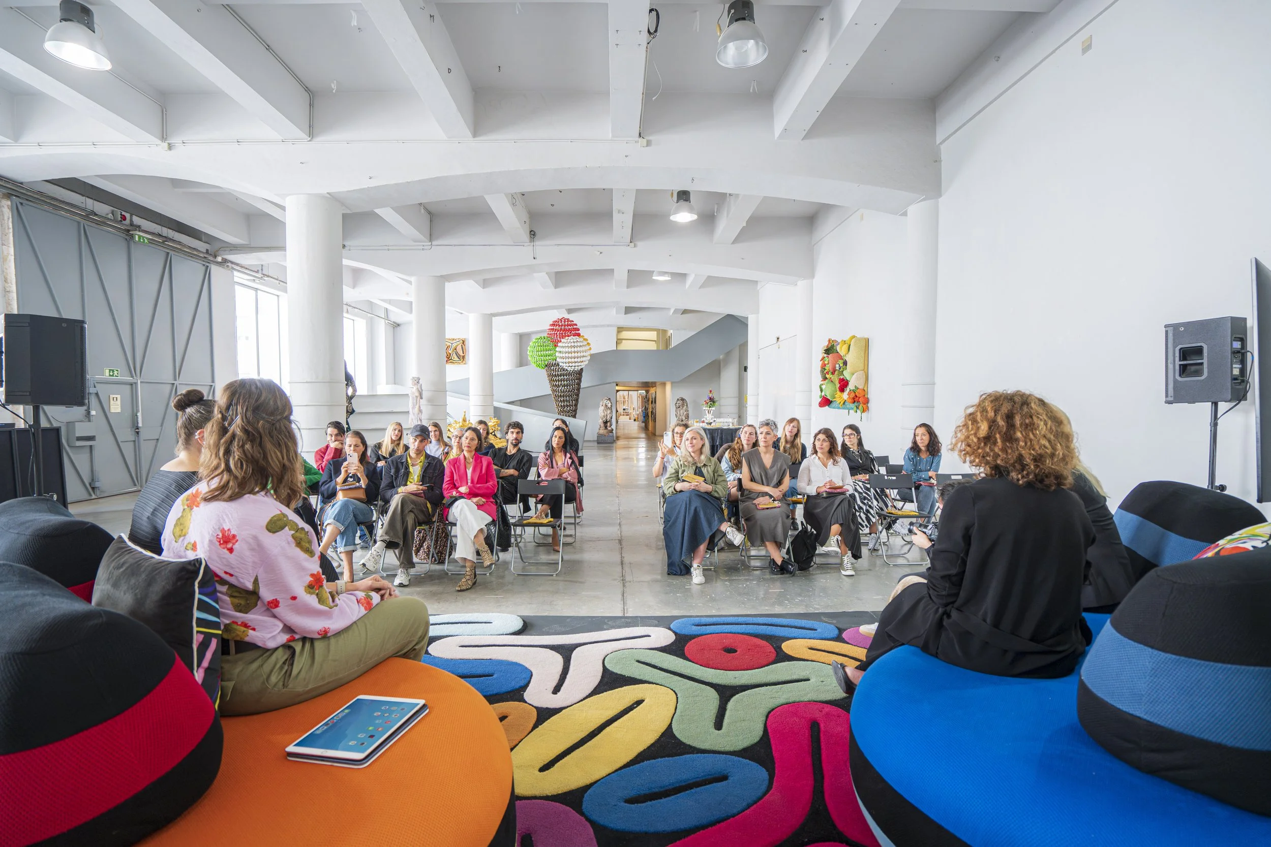 Pessoas sentadas em cadeiras e almofadas em um evento ou reunião em um espaço amplo, com paredes brancas e decoração colorida, incluindo uma escultura de bolas e obras de arte nas paredes.