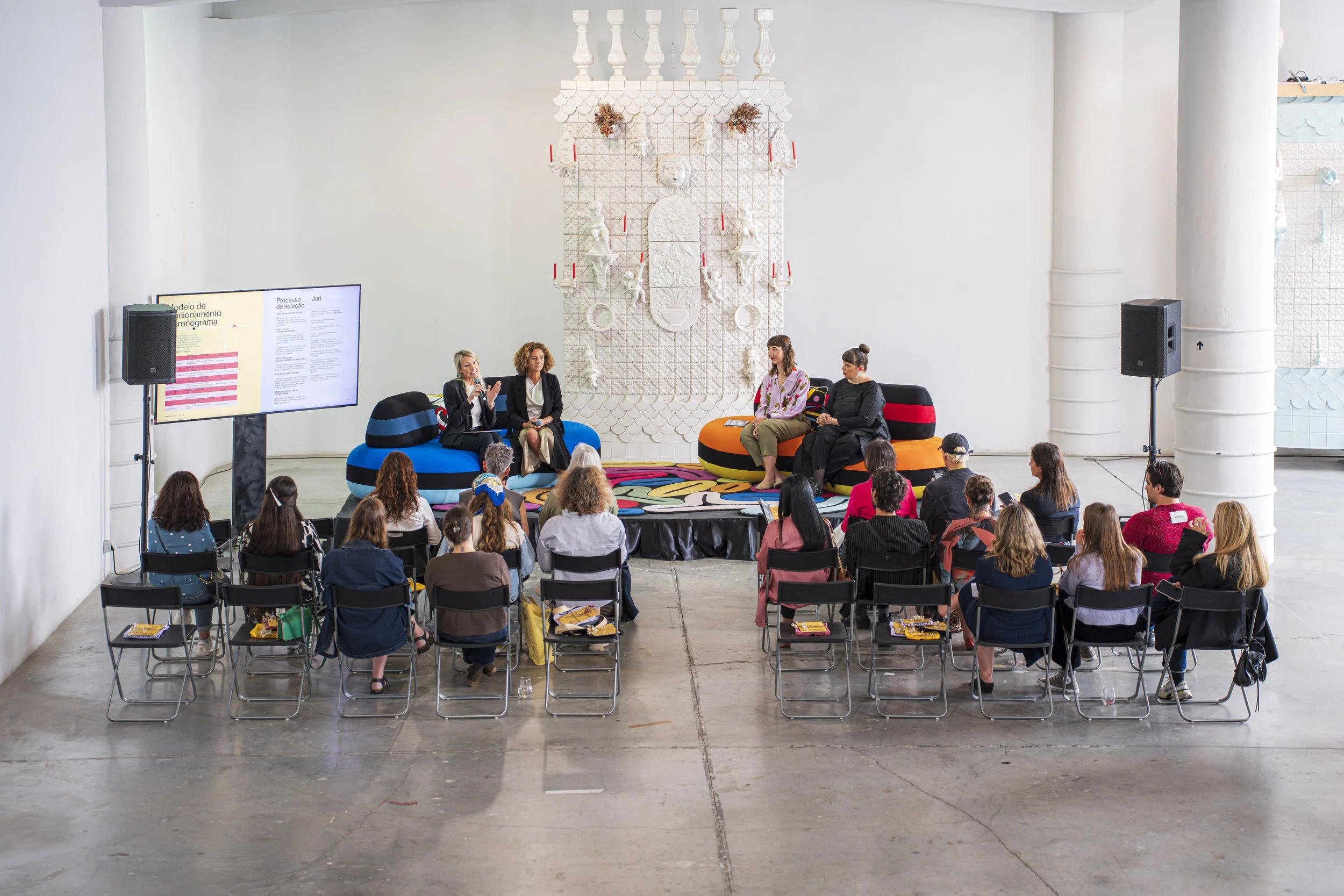 Palestrantes participando de uma mesa-redonda em um evento, sentados em poltronas coloridas, com uma grande instalação de decoração branca ao fundo e uma tela de projeção ao lado esquerdo.