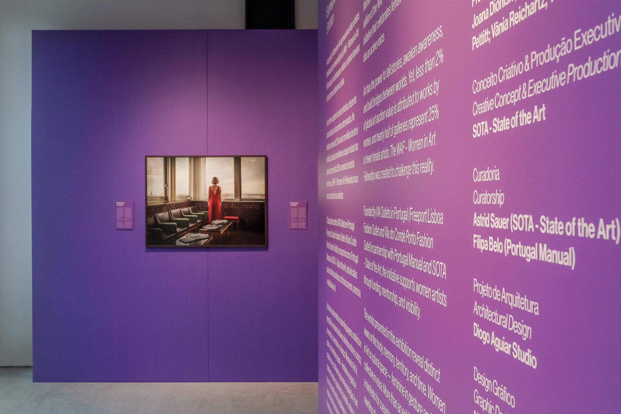Mural roxo com texto em inglês e português, explicando o conceito de 'Woman in Art' e os detalhes de uma exposição artística, com uma TV que mostra uma mulher de vestido vermelho na sala.
